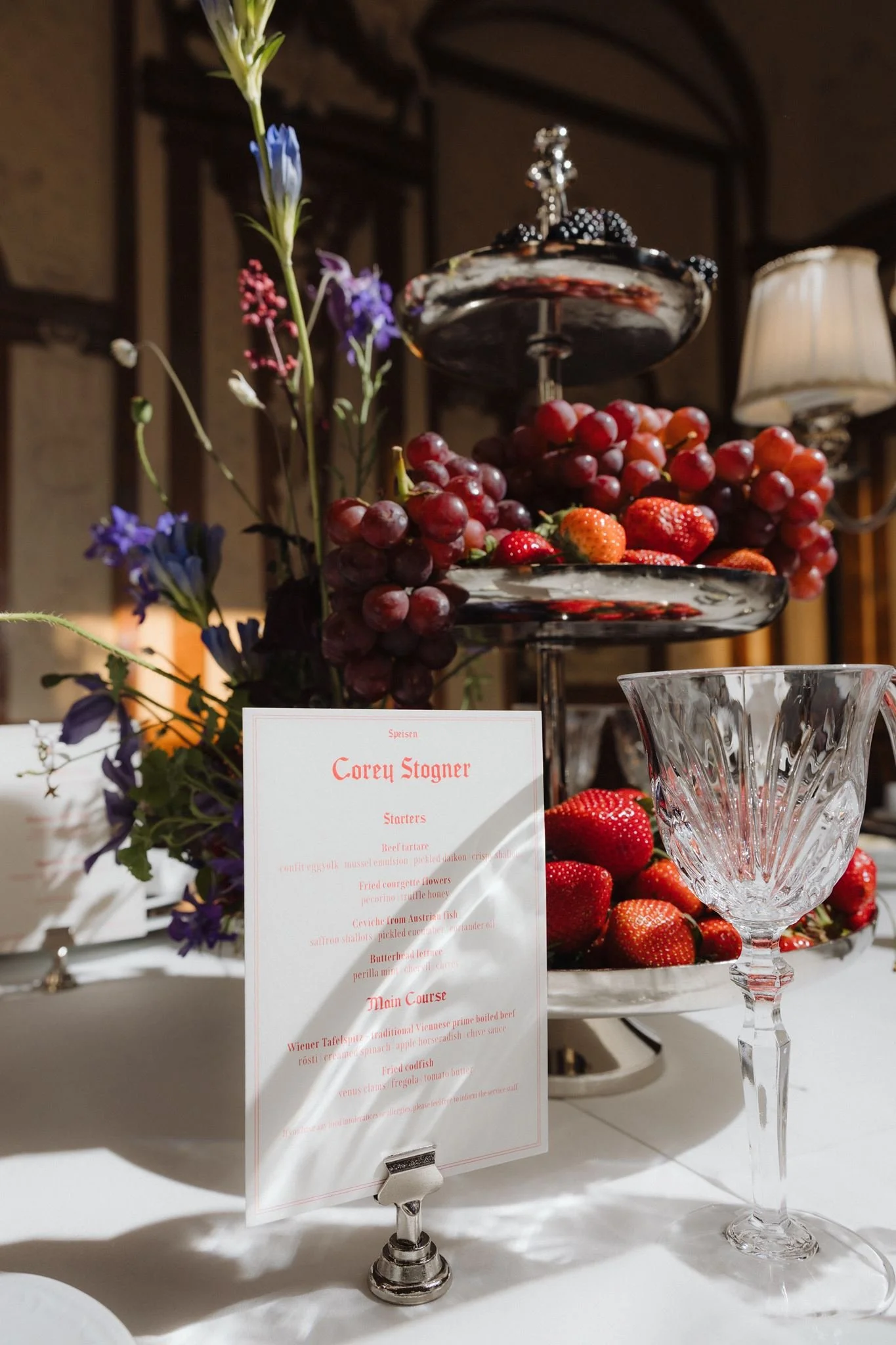 A tiered dessert stand filled with strawberries and grapes, decorated with purple and red flowers, placed on a white table with a menu card in front.