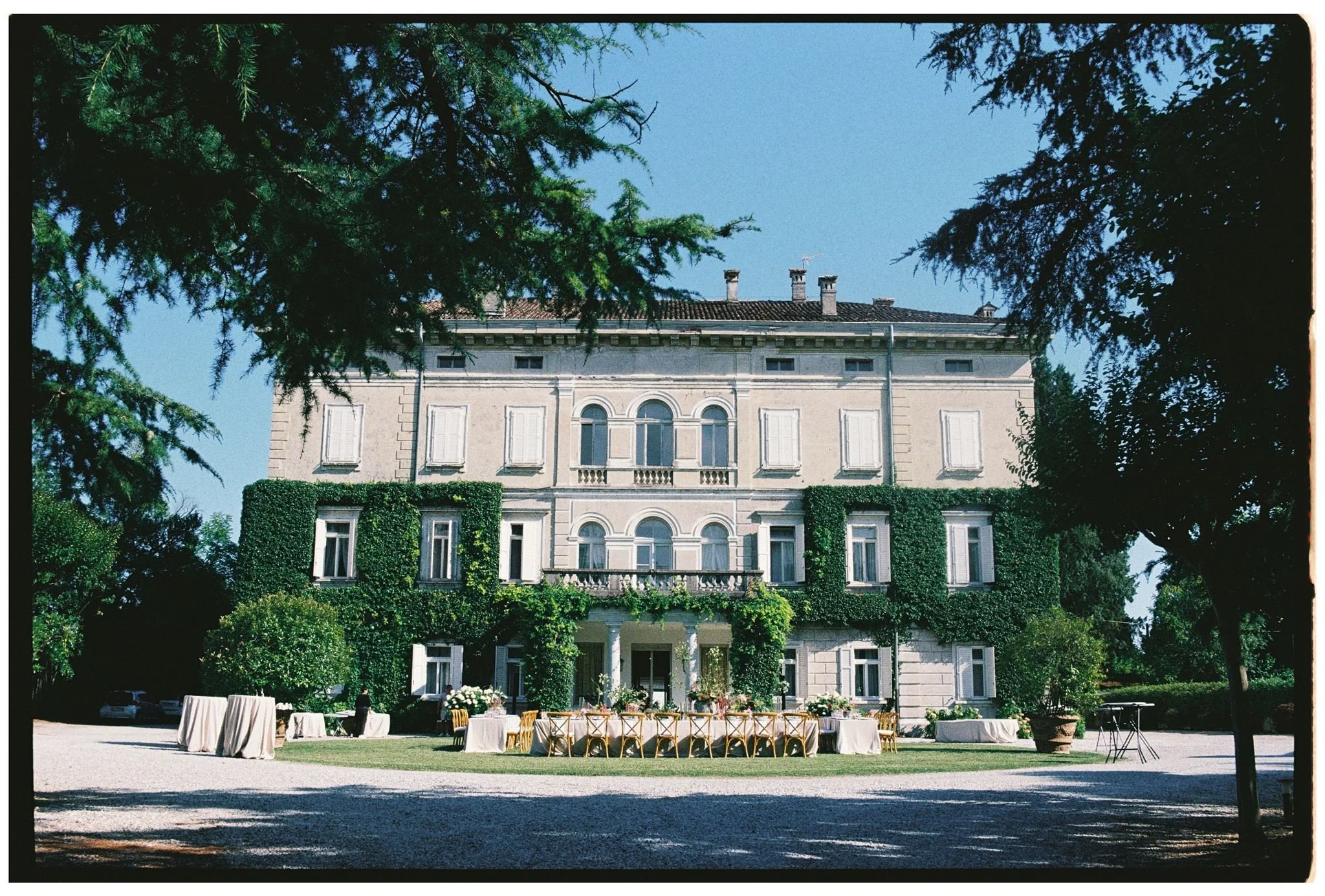 Elegant multi-story mansion surrounded by lush greenery, with tables and chairs set up for an outdoor event.