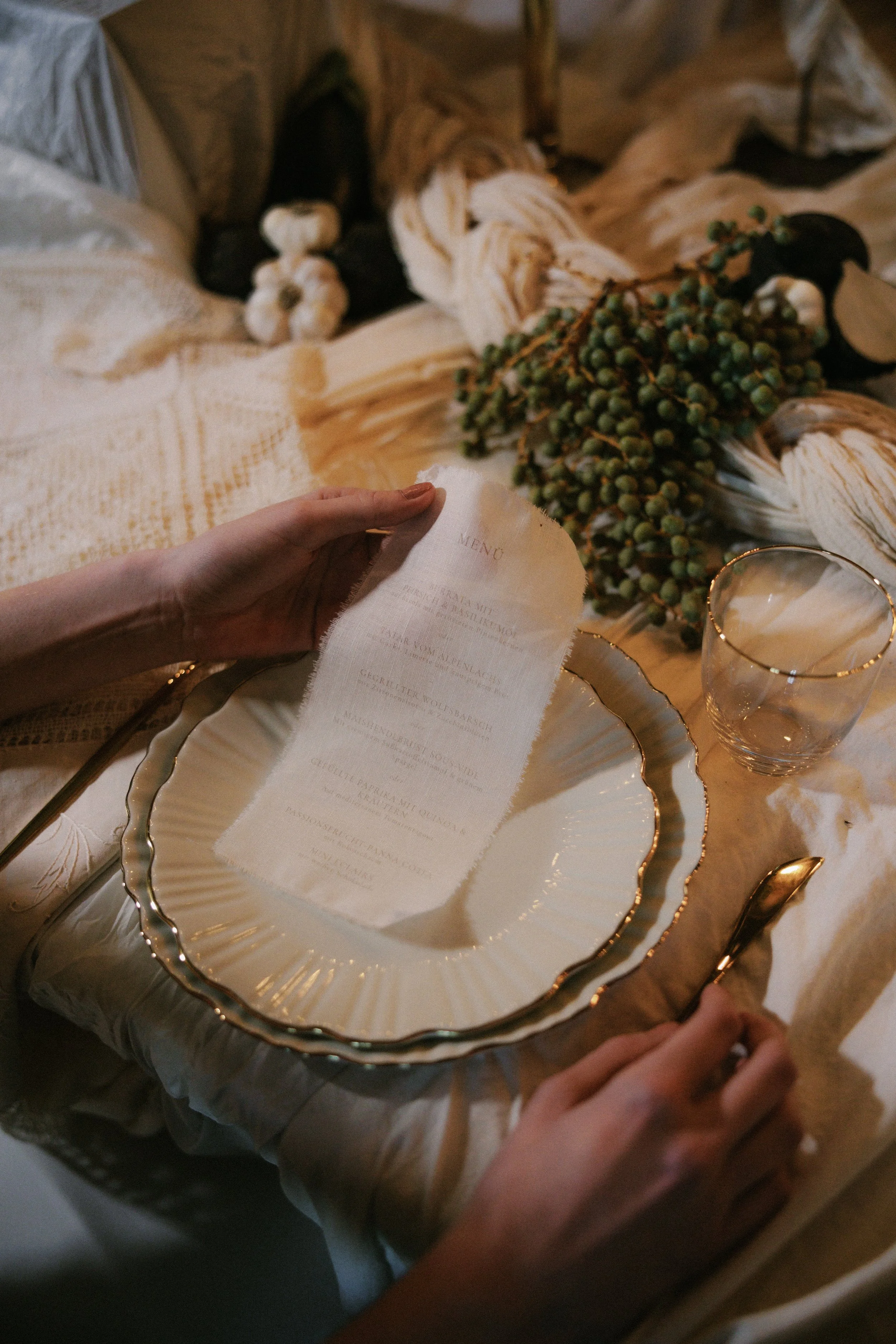 Person holding a torn paper menu above a decorative plate on a table with a glass of water, cloth, and a bunch of green berries or grapes.
