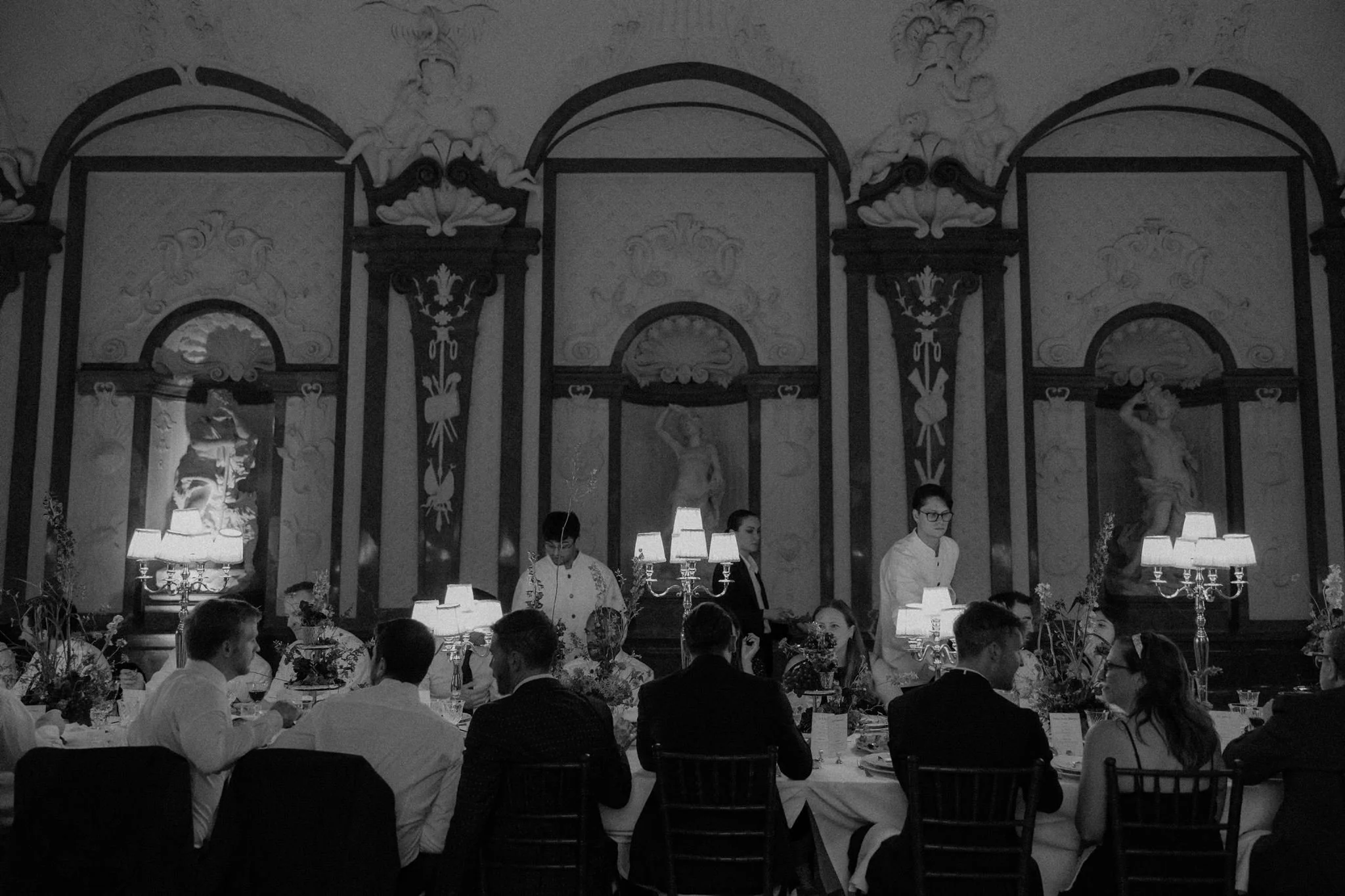 Elegant dining room with ornate wall decorations, multiple table lamps, and waitstaff serving guests at a formal event.