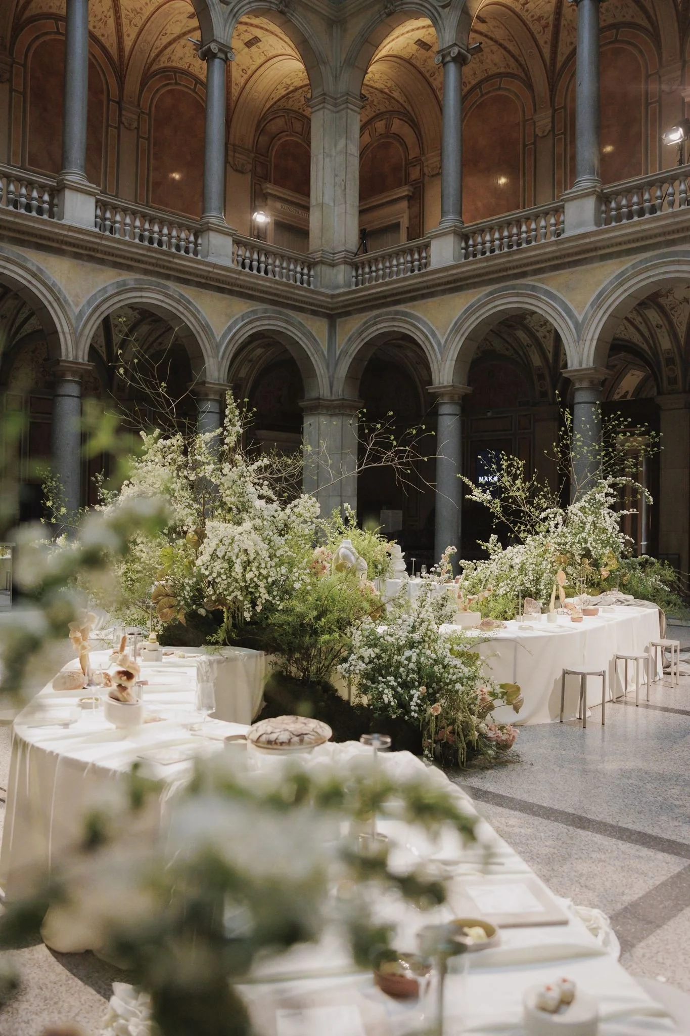 Elegant indoor event space with multiple levels, ornate arches, and columns, decorated with fresh white and light pink flowers on tables.