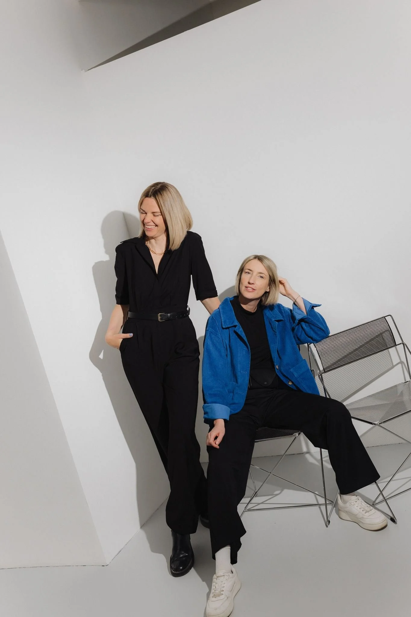 Two women with blonde hair in casual attire, one standing in black and the other sitting in blue, sharing a moment in a minimalist room with white walls and a gray floor. They appear to be enjoying themselves.