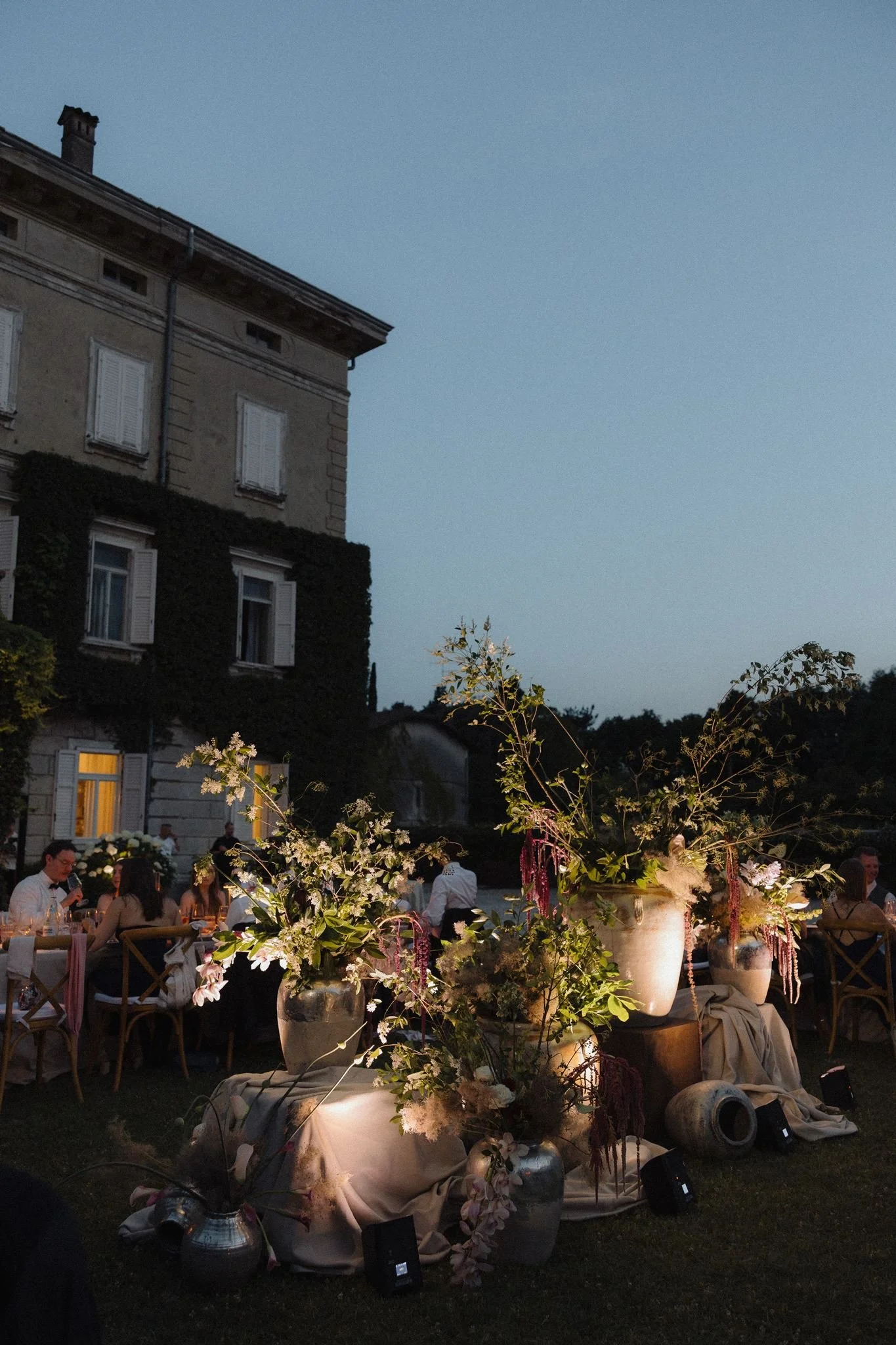 A group of people dining outdoors at an evening event, with a large floral centerpiece in the foreground, surrounded by decorative vases and candles, against a backdrop of a building with windows and a clear evening sky.