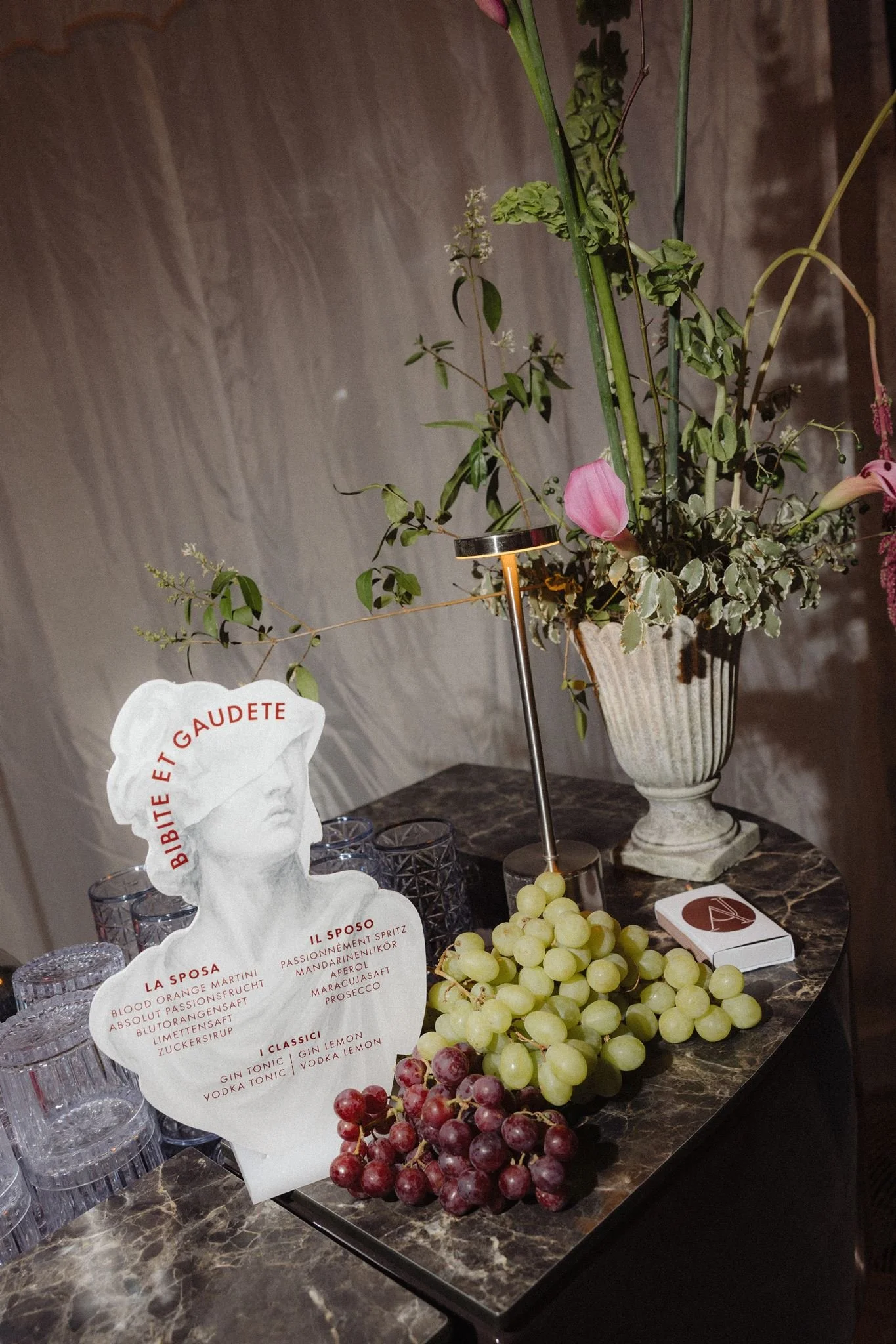 A floral arrangement with pink lilies and greenery in a white vase, set on a dark marble table, surrounded by grapes and glassware, with a menu and a decorative paper with a face silhouette.