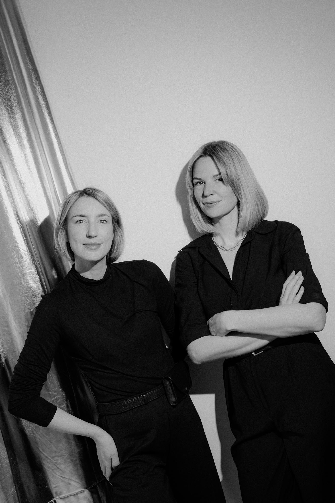 Two women standing against a plain wall, both wearing black outfits and smiling at the camera.