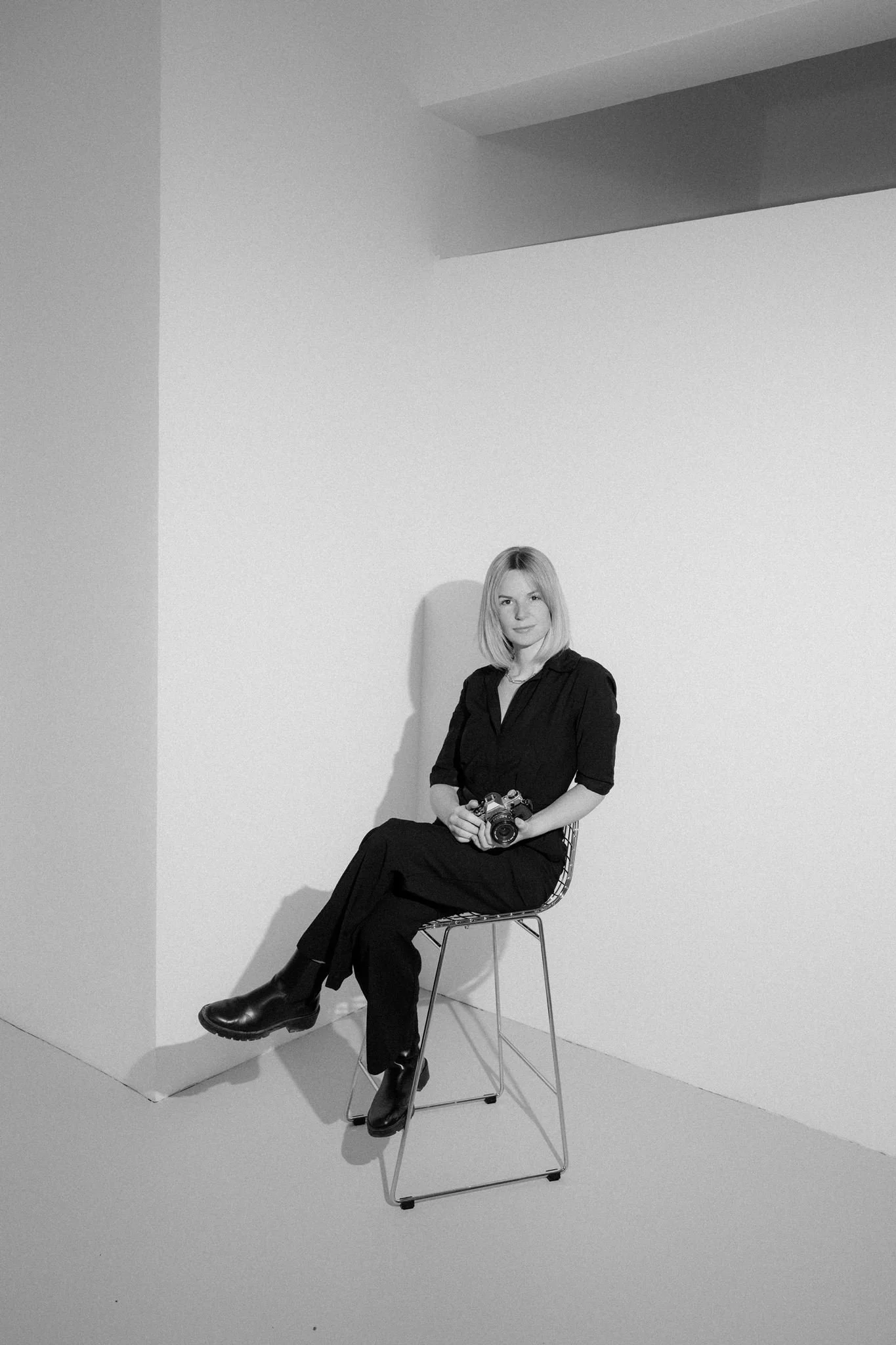 A woman with blonde hair, wearing a black outfit and black boots, sitting sideways on a metal chair, holding a camera, in a minimalistic room with white walls and a coffered ceiling.
