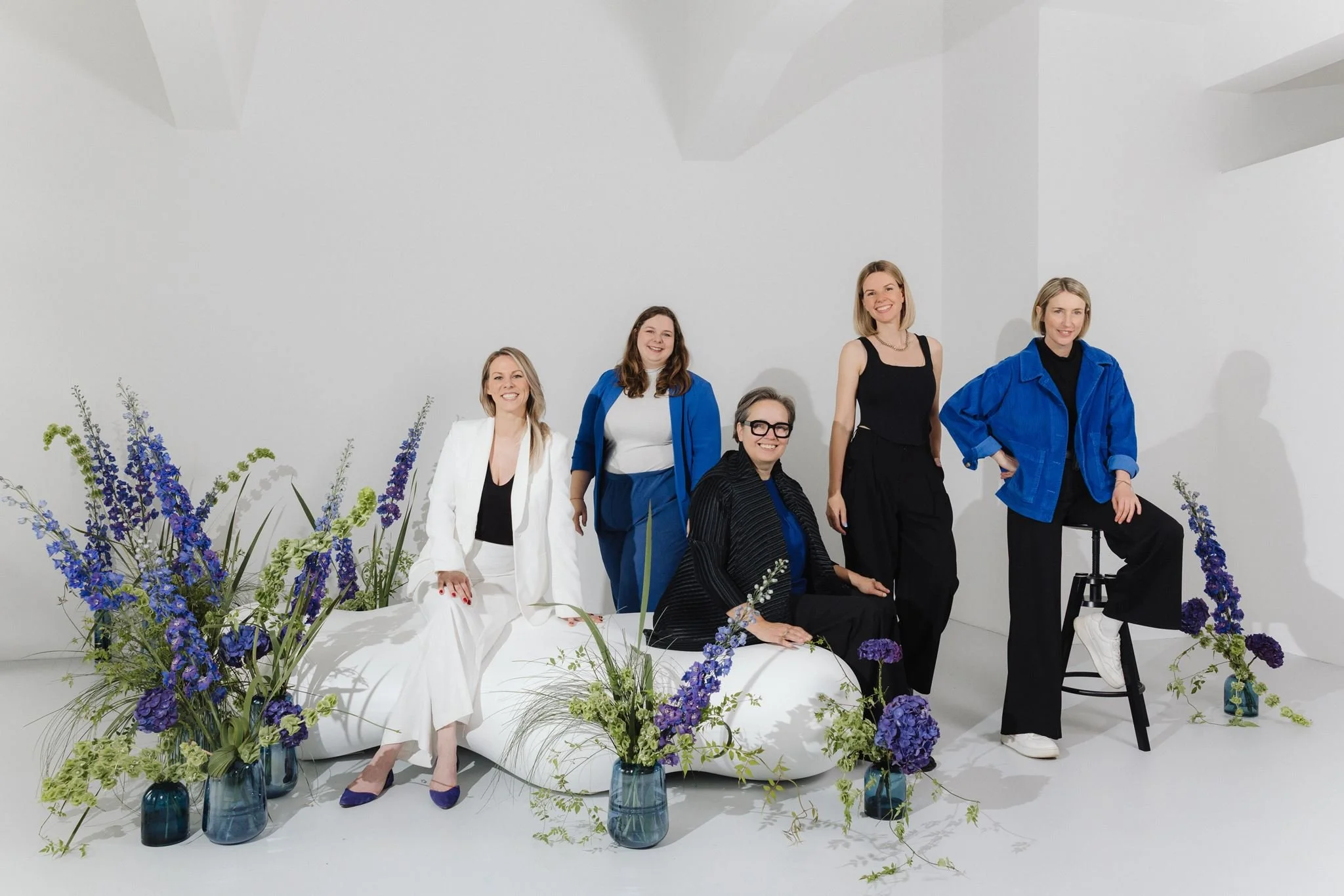 Six women with blond and brunette hair, dressed in stylish outfits, posing for a photo in a white studio setting surrounded by purple and green floral arrangements in blue vases.