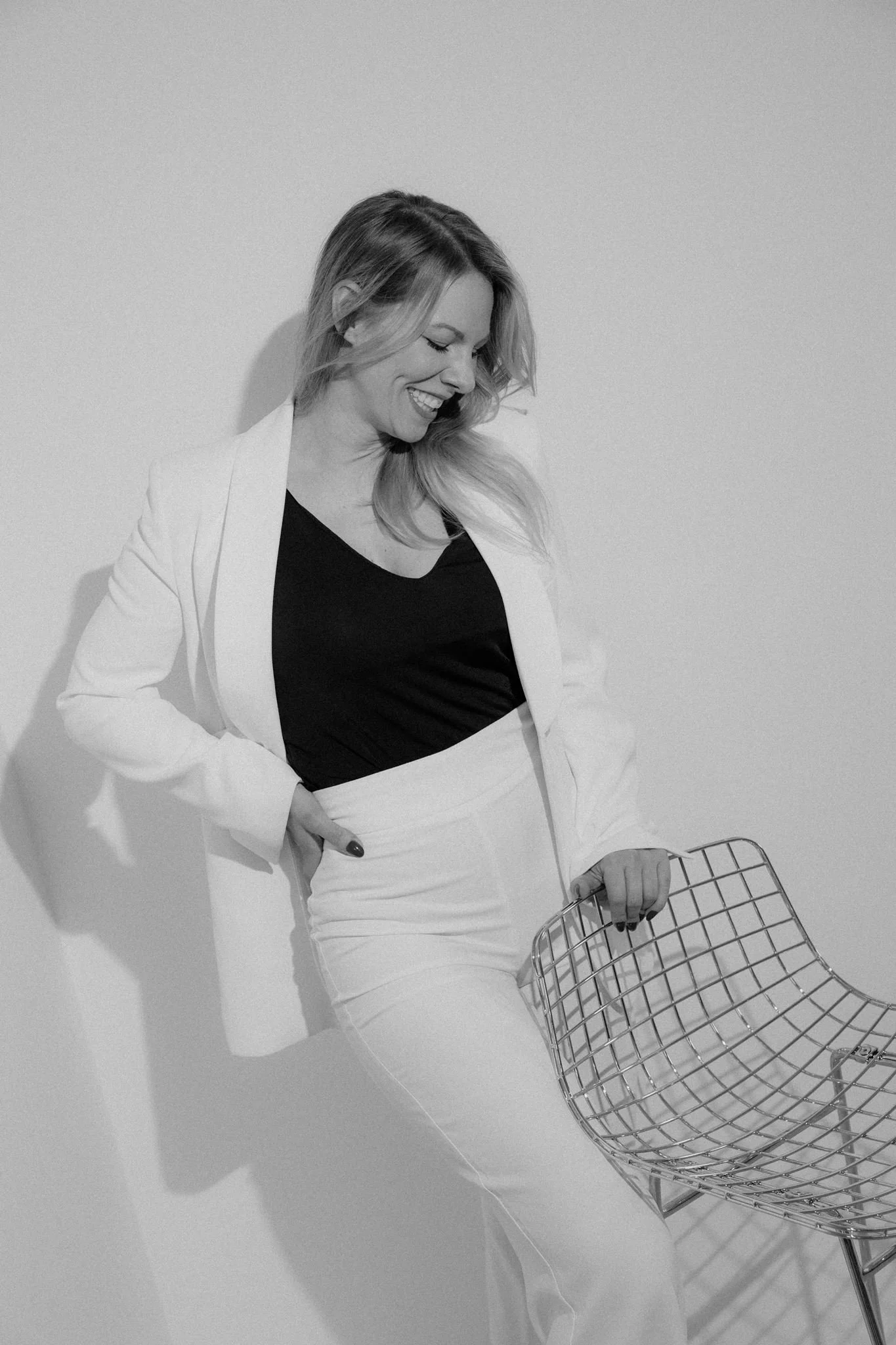 A woman with long hair smiling and looking down, wearing a blazer and white pants, holding a shopping cart against a plain wall.