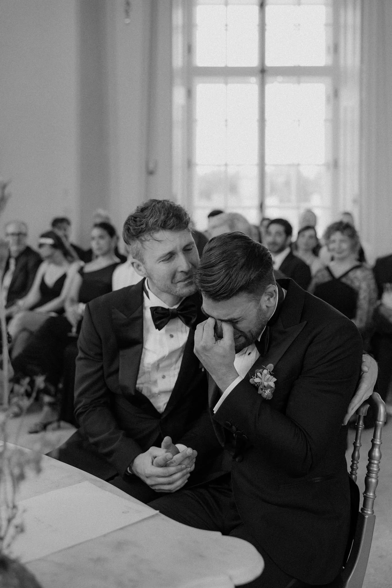 A wedding reception with two men in tuxedos, one of whom is emotional, holding hands and sitting close in front of a large window, while guests are seated in the background.