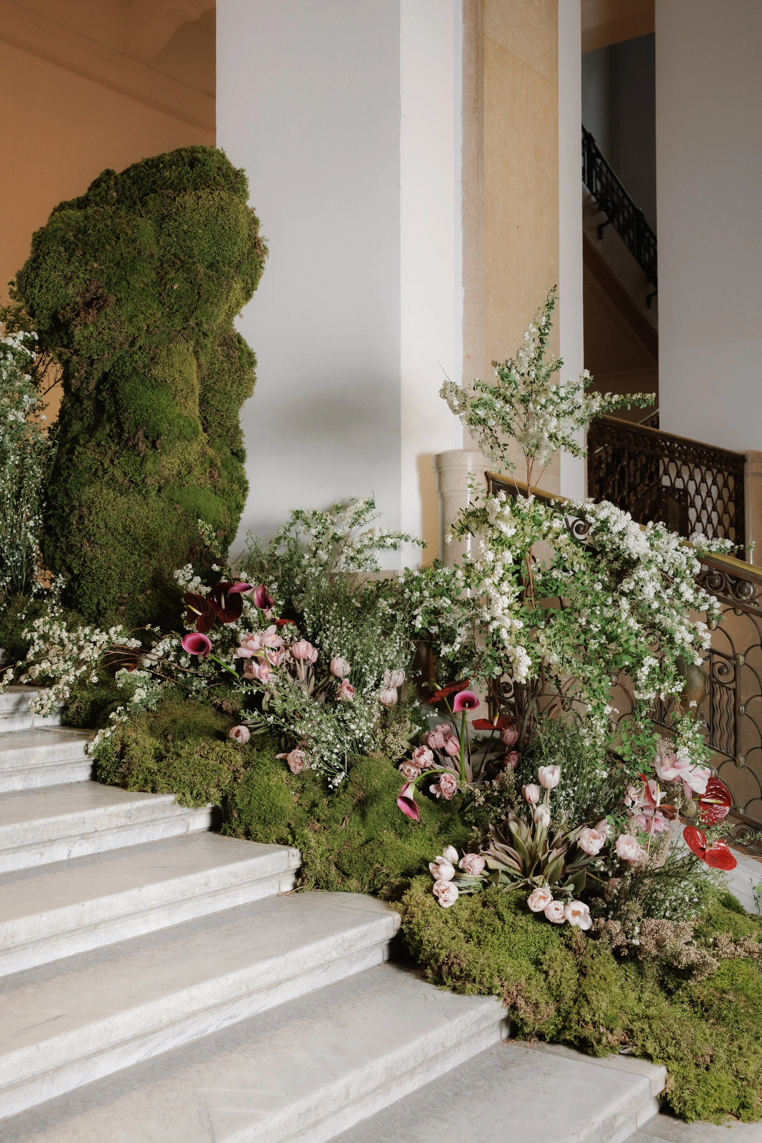 A staircase decorated with various flowers and greenery, including white blooms, pink tulips, calla lilies, and lush bushes, next to an elegant wrought iron handrail in a well-lit indoor space.