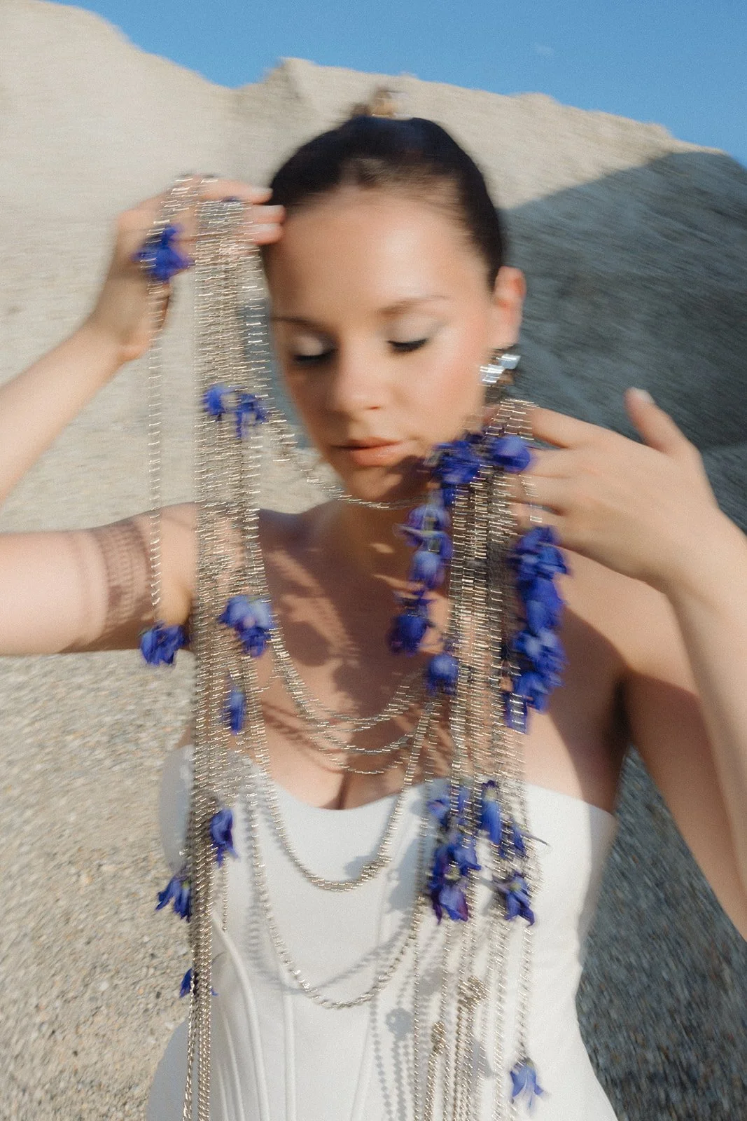 A woman with dark hair styled in an updo, wearing a white strapless dress, is adorned with multiple layered silver chains and blue flower accessories. She is holding some of the chains with both hands, and her eyes are closed. The background features a rocky, outdoor landscape with a blue sky.
