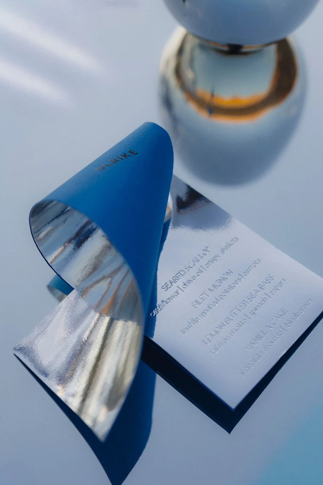 A blue and silver knife with the word 'UNLIKELY' engraved on it, resting on a menu or list, with a glass and a metallic object reflecting in the background.