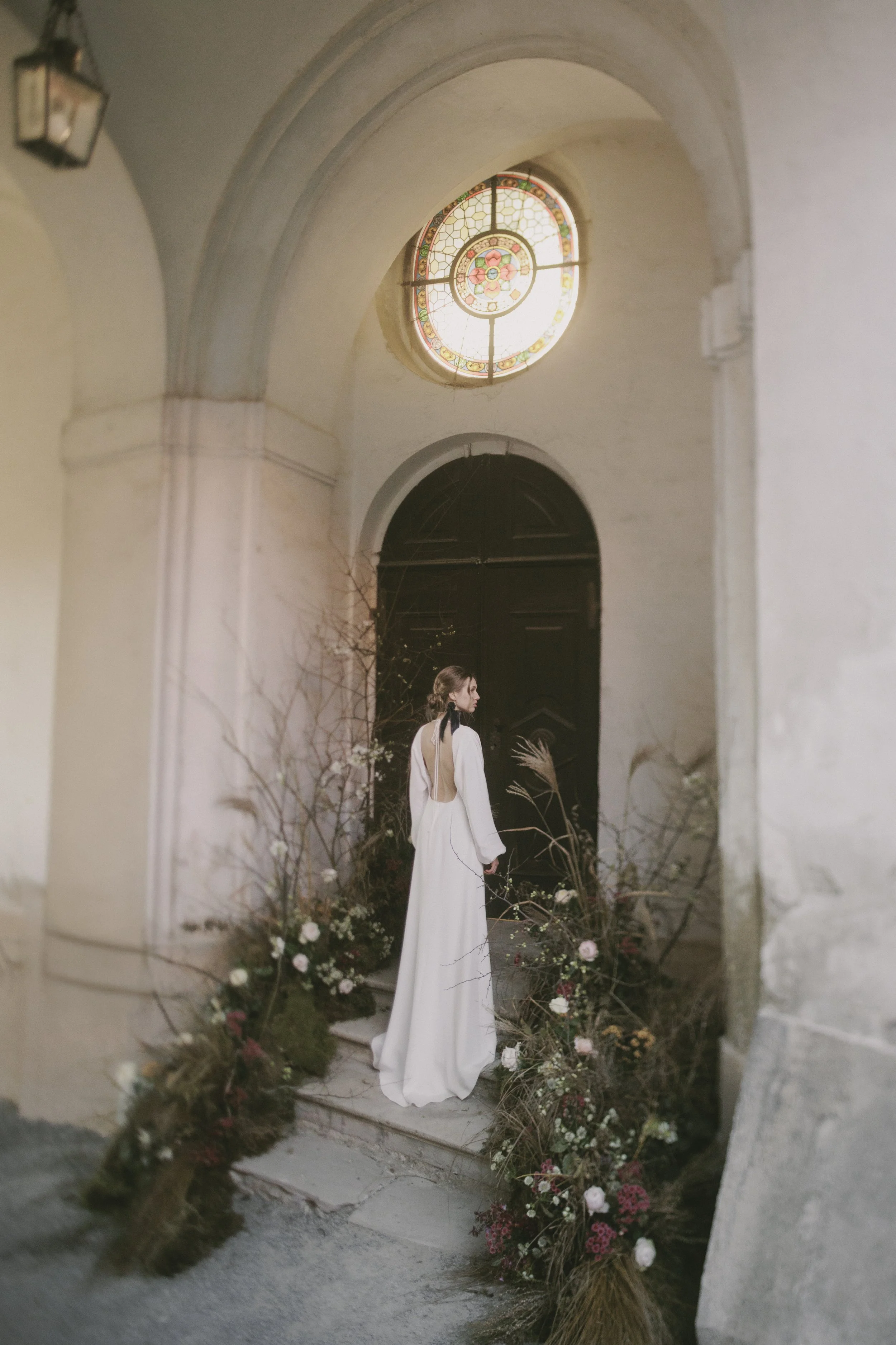 A woman in a long white dress with an open back standing in front of a dark door, surrounded by flowers and branches, inside a building with an arched ceiling and a stained-glass window.