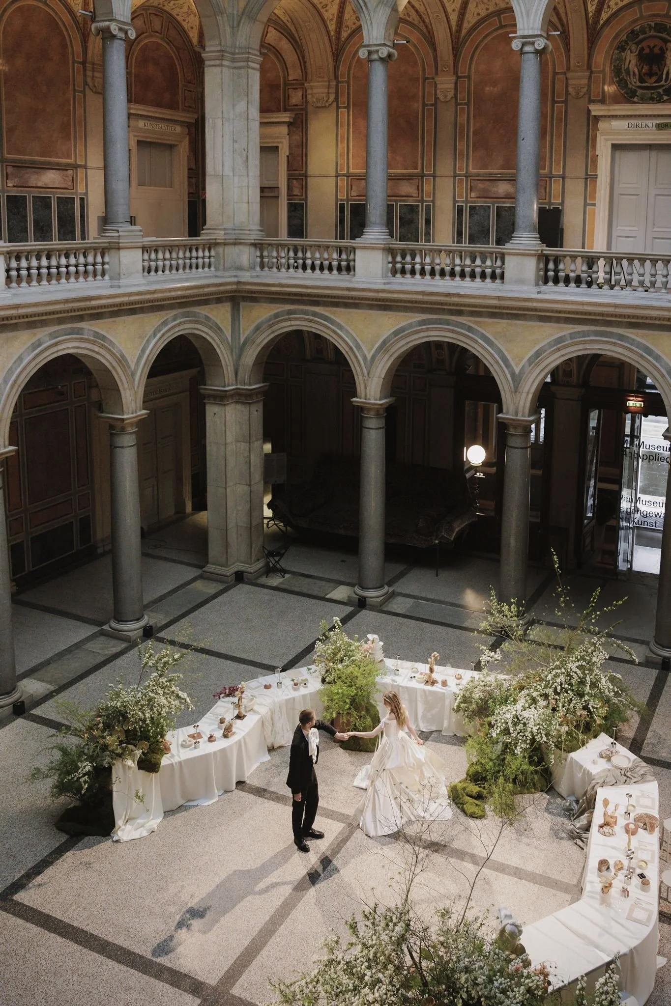 A wedding reception with a couple dancing in a grand, historic hall with arches, columns, and ornate decorations, surrounded by tables with desserts and floral arrangements.