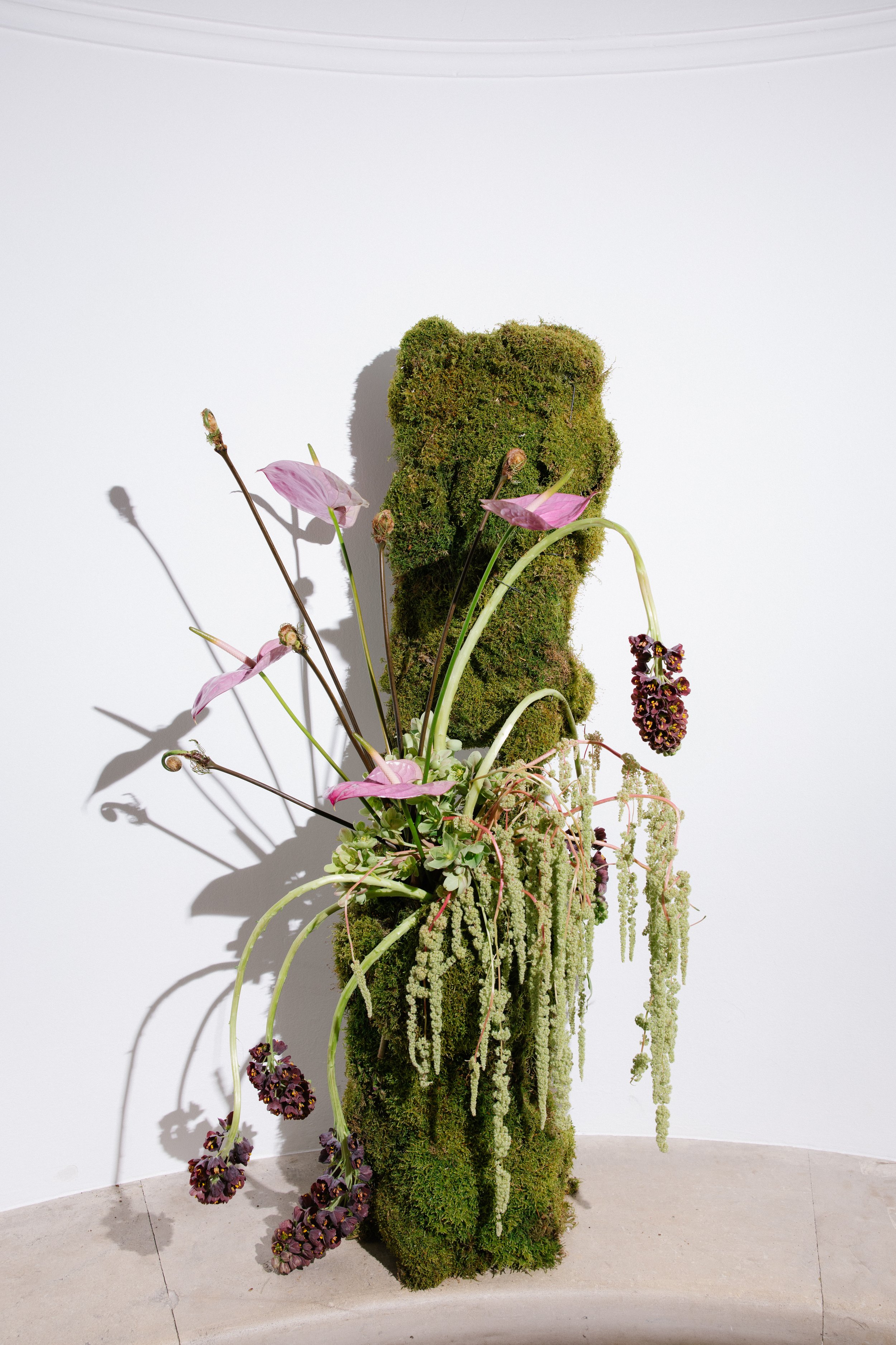 A tall sculpture made of moss and flowers, resembling a bear, stands against a white wall. The base is covered in moss, with pink and dark purple flowers arranged around and on top, creating an artistic floral display.