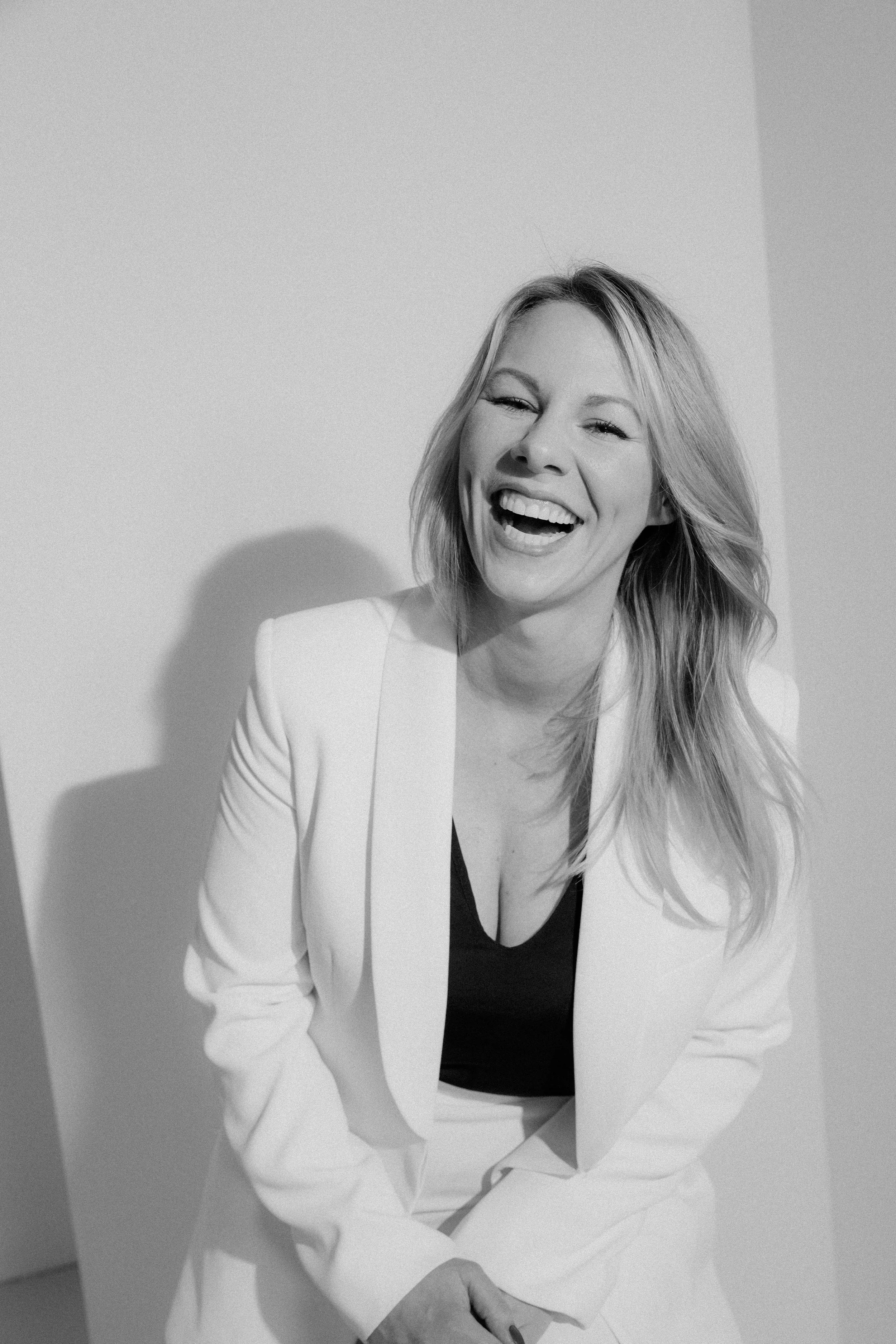 Black and white photo of a woman wearing a white blazer and black top, smiling and winking, with her hair styled in loose waves, sitting against a plain wall.