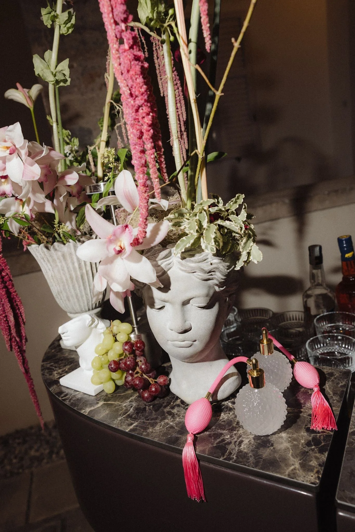 A decorative table display featuring a white head-shaped vase with closed eyes, filled with pink and white orchids, purple and pink amaranthus, and variegated foliage; also includes clusters of green and red grapes, small glass containers, and pink tassel and jewel-shaped accessories.
