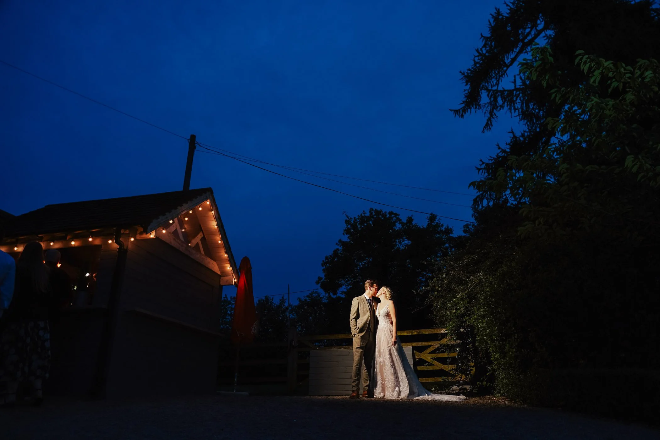 A bride and groom standing closely together outdoors during their wedding at night, under a dark blue sky, with trees and string lights in the background.