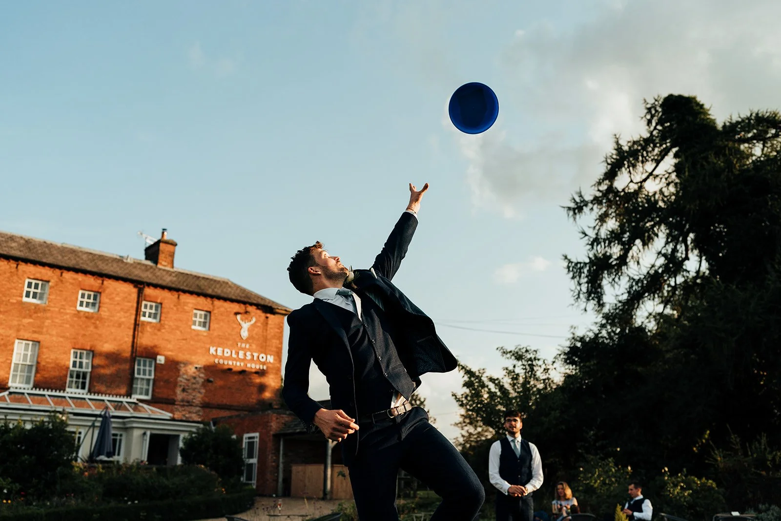 A man in a suit throwing a blue hat into the air outside near a brick building with a sign that reads 'Kedleton Country House' and some other people in the background.