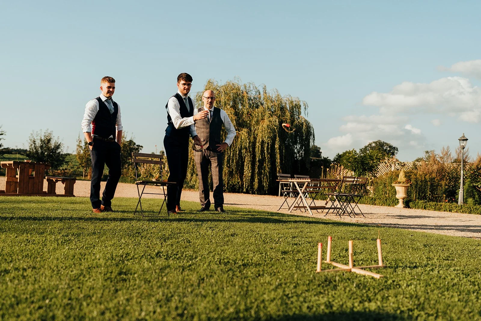 Three men in formal attire playing cornhole outdoors on a lawn with tables, chairs, and a decorative garden in the background.