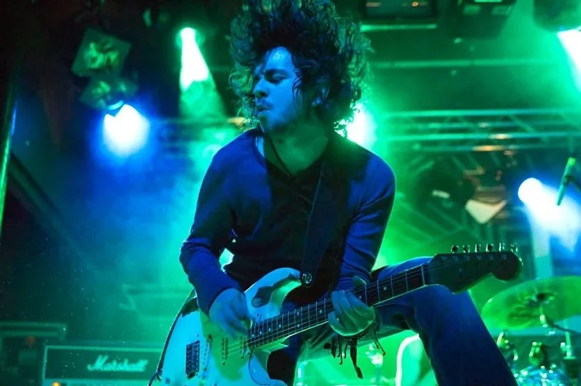 A musician with curly hair playing an electric guitar on stage with green and blue lighting.