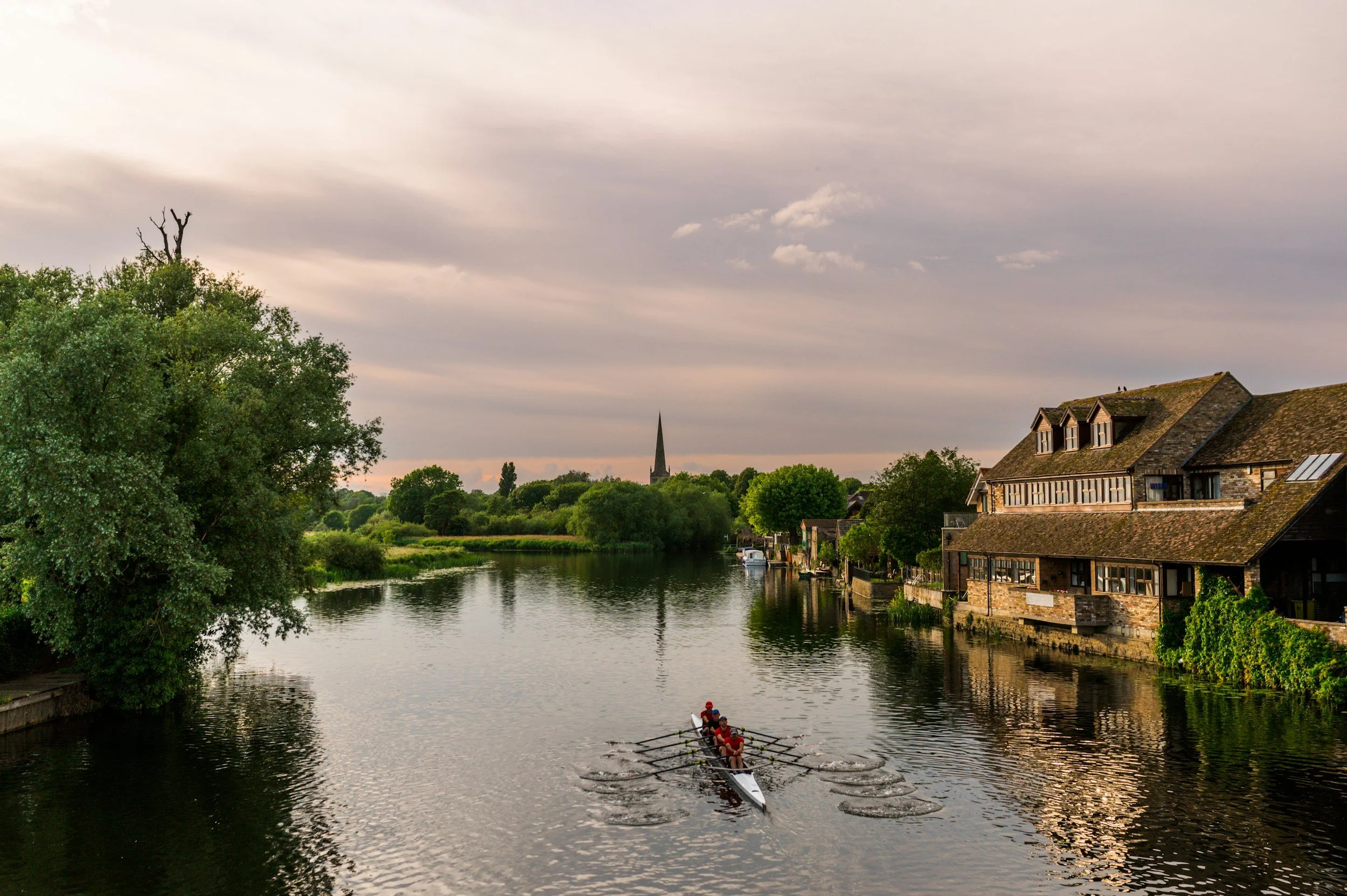 A scenic river scene with a rowboat carrying five people, houses along the riverbank, lush green trees, and a church steeple in the distance under a cloudy sky. This is St Ives, Cambridgeshire.