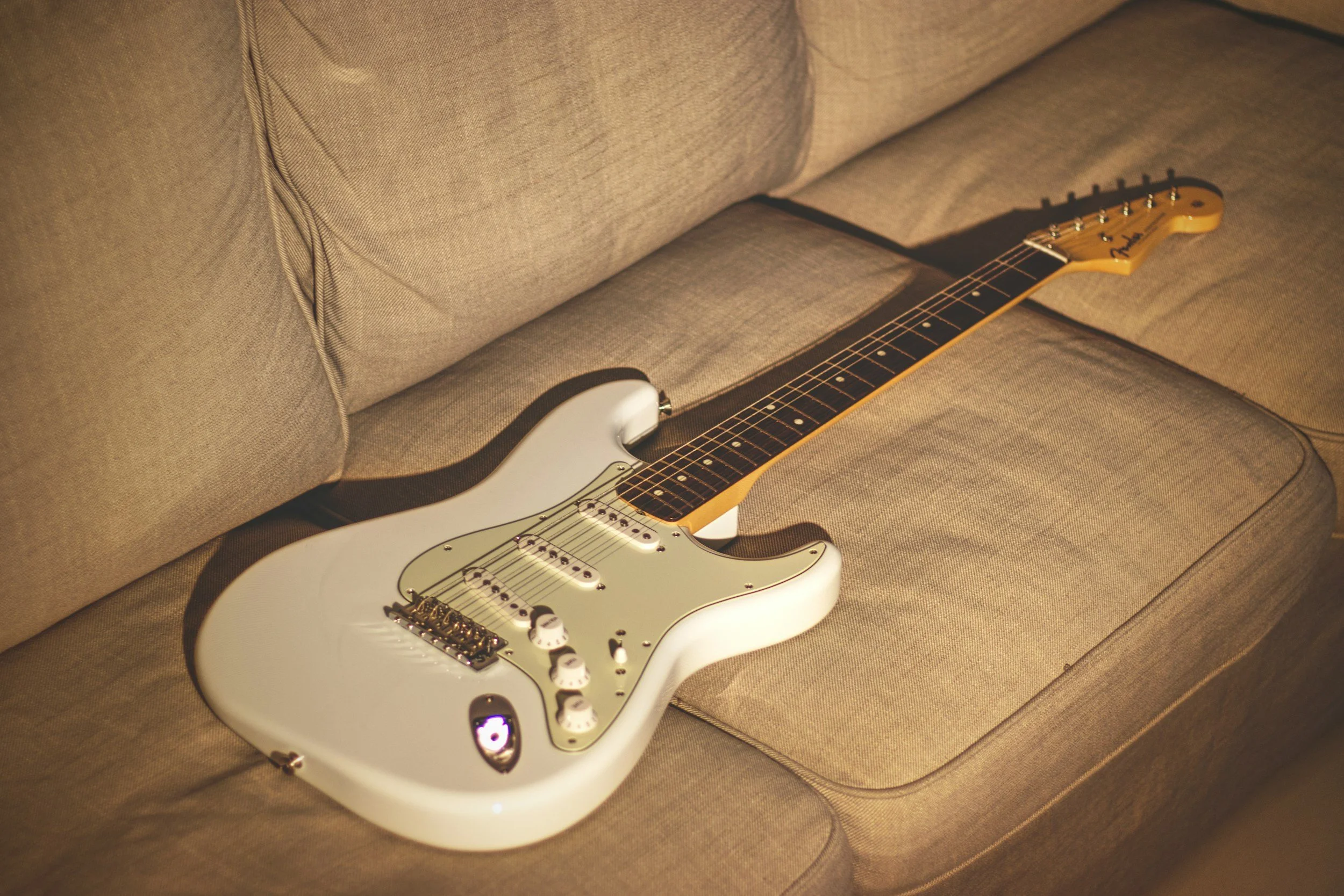 A white electric guitar resting on a tan fabric sofa.