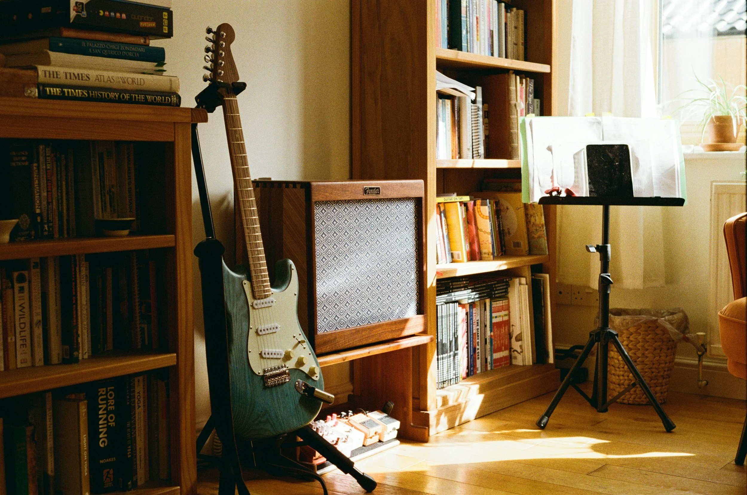 A room with bookshelves filled with books, a partially visible electric guitar leaning against a bookshelf, a small amplifier, a music stand, and a window with sunlight coming through. A potted plant is on the windowsill.
