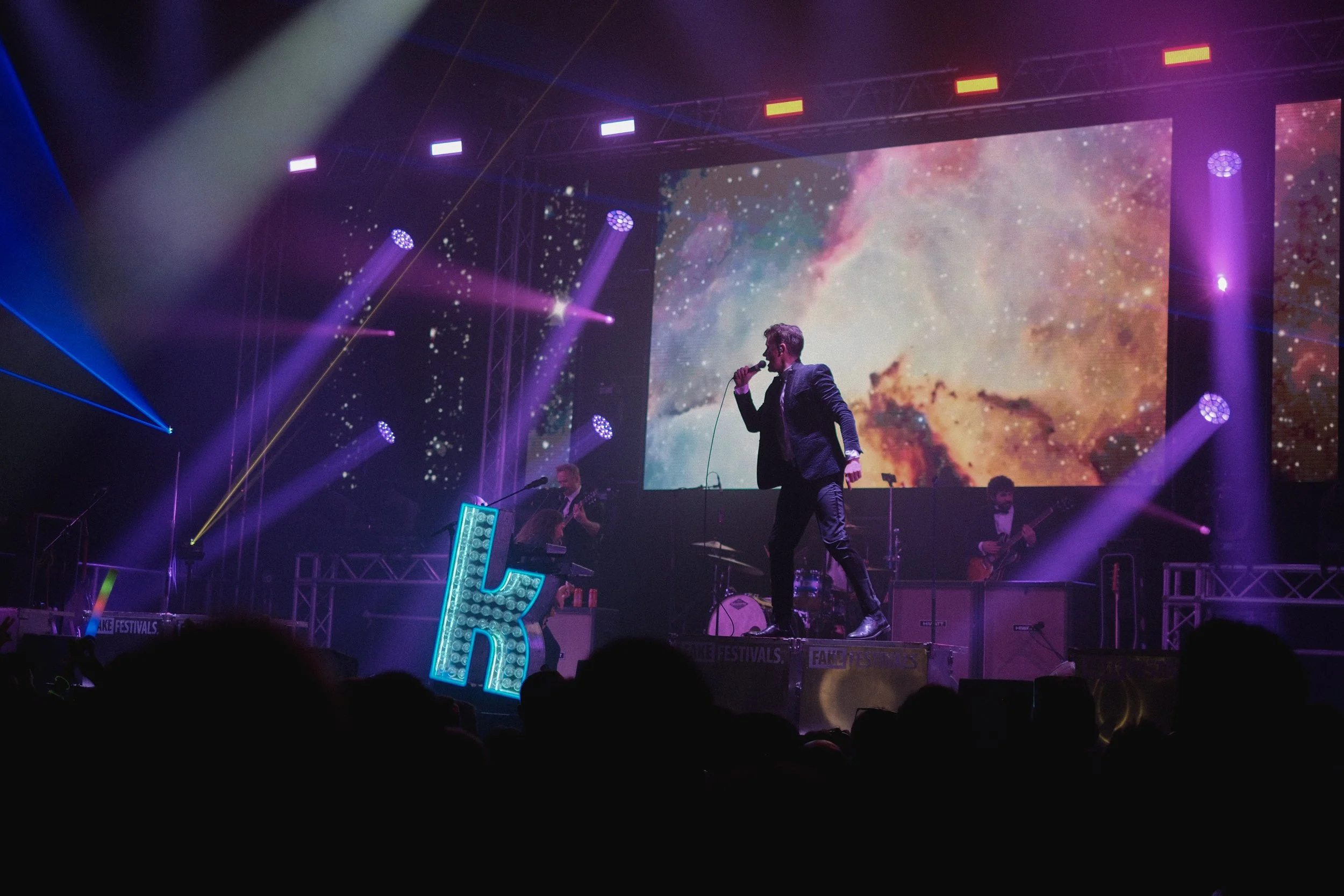 A live music performance on a stage with a large galaxy-themed display in the background, colorful stage lighting, and a singer holding a microphone. There are band members playing instruments and a large illuminated letter 'B' on the stage.