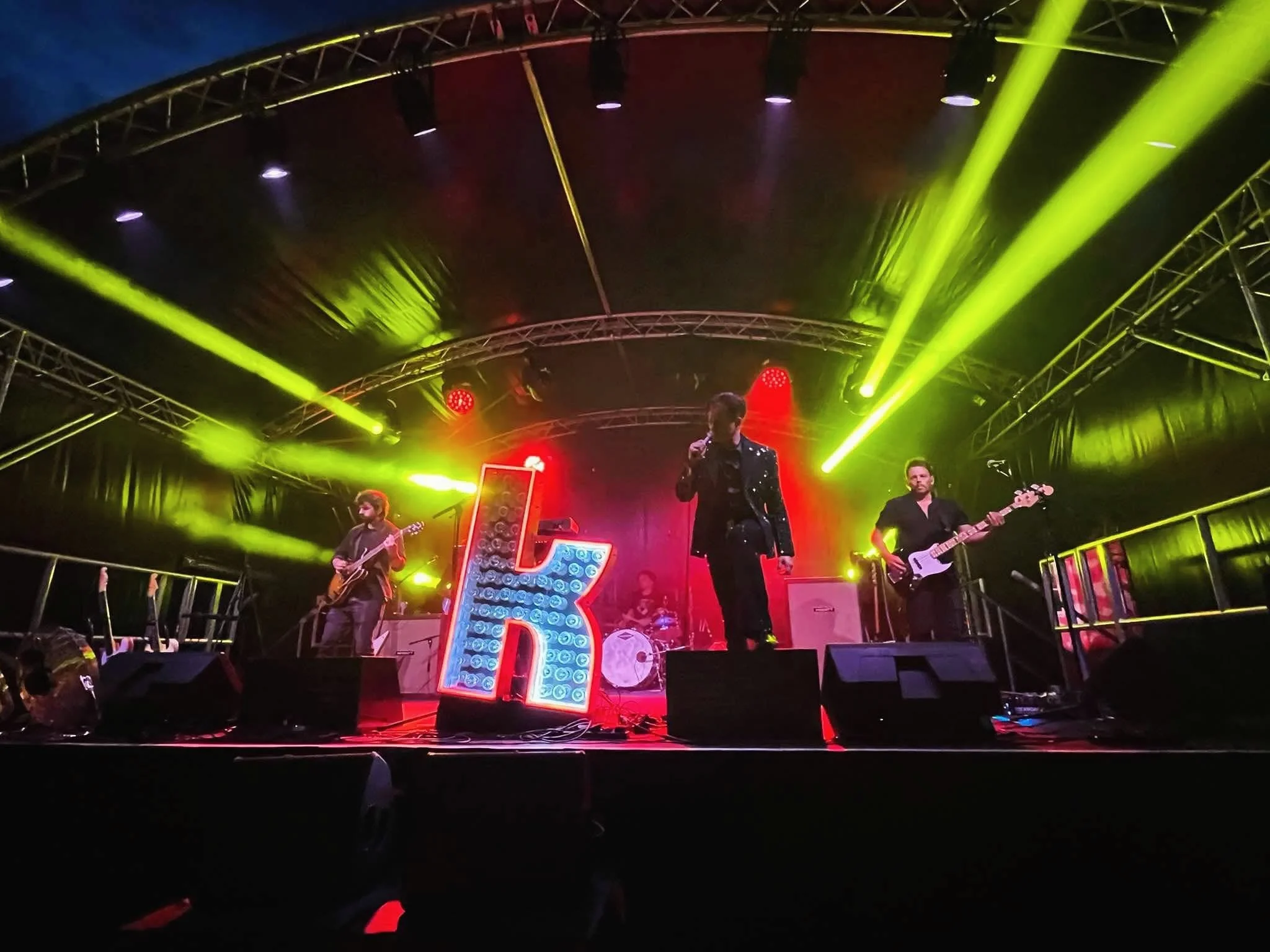 A live music performance on a stage with bright yellow and purple stage lights, featuring a band with several musicians playing instruments and singing.