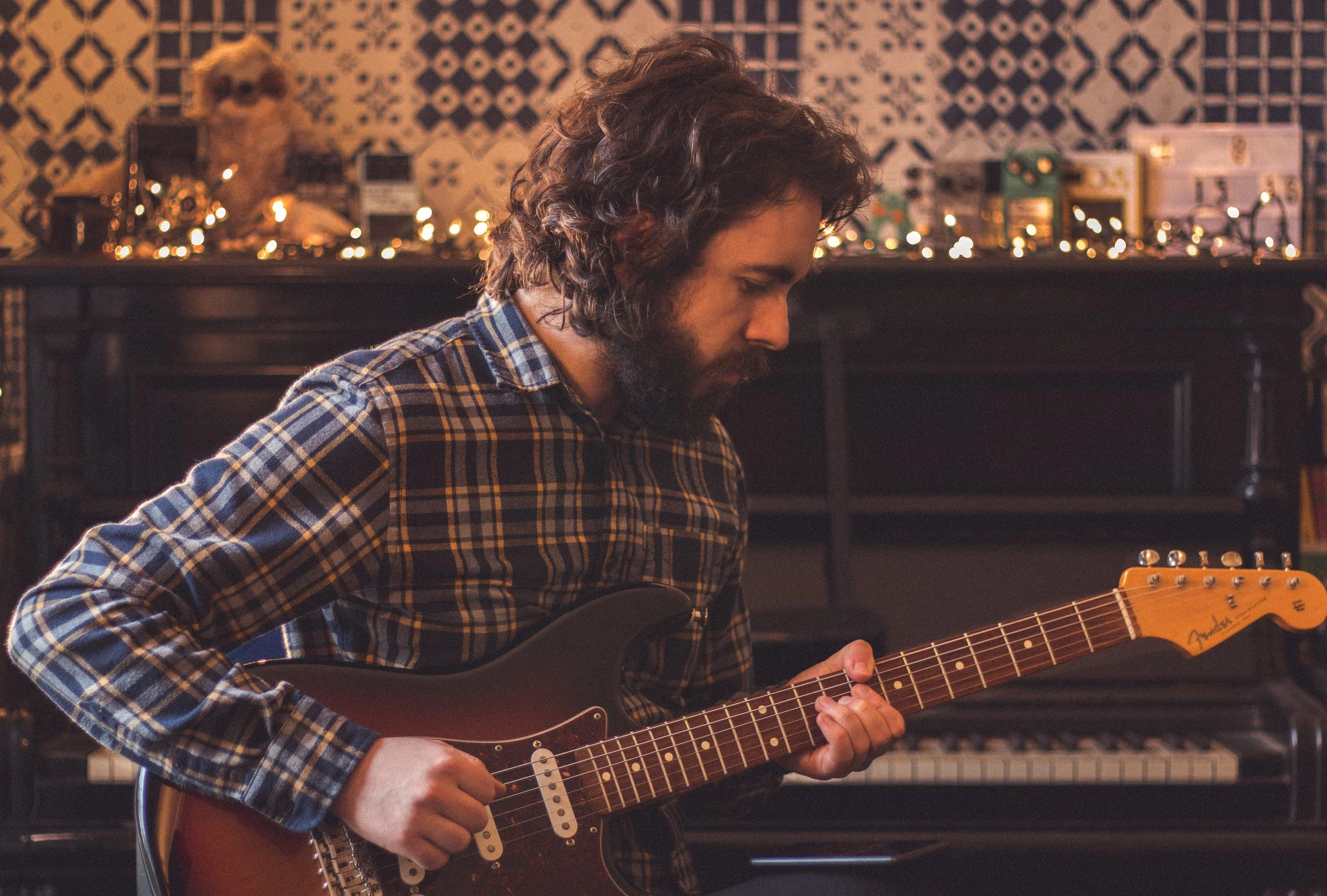 A man with curly hair and a beard wearing a plaid shirt, playing an electric guitar in a room with musical equipment and string lights in the background.