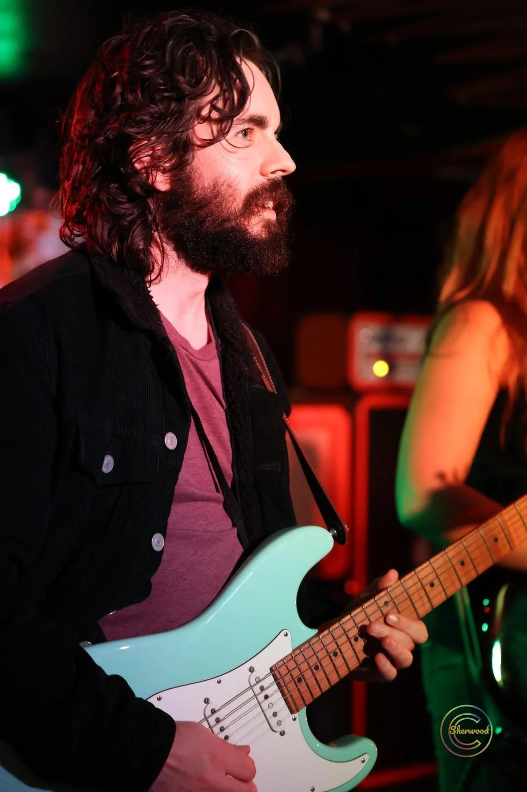 A man with long dark curly hair and a beard playing an electric guitar in a dimly lit venue, with colorful stage lighting.