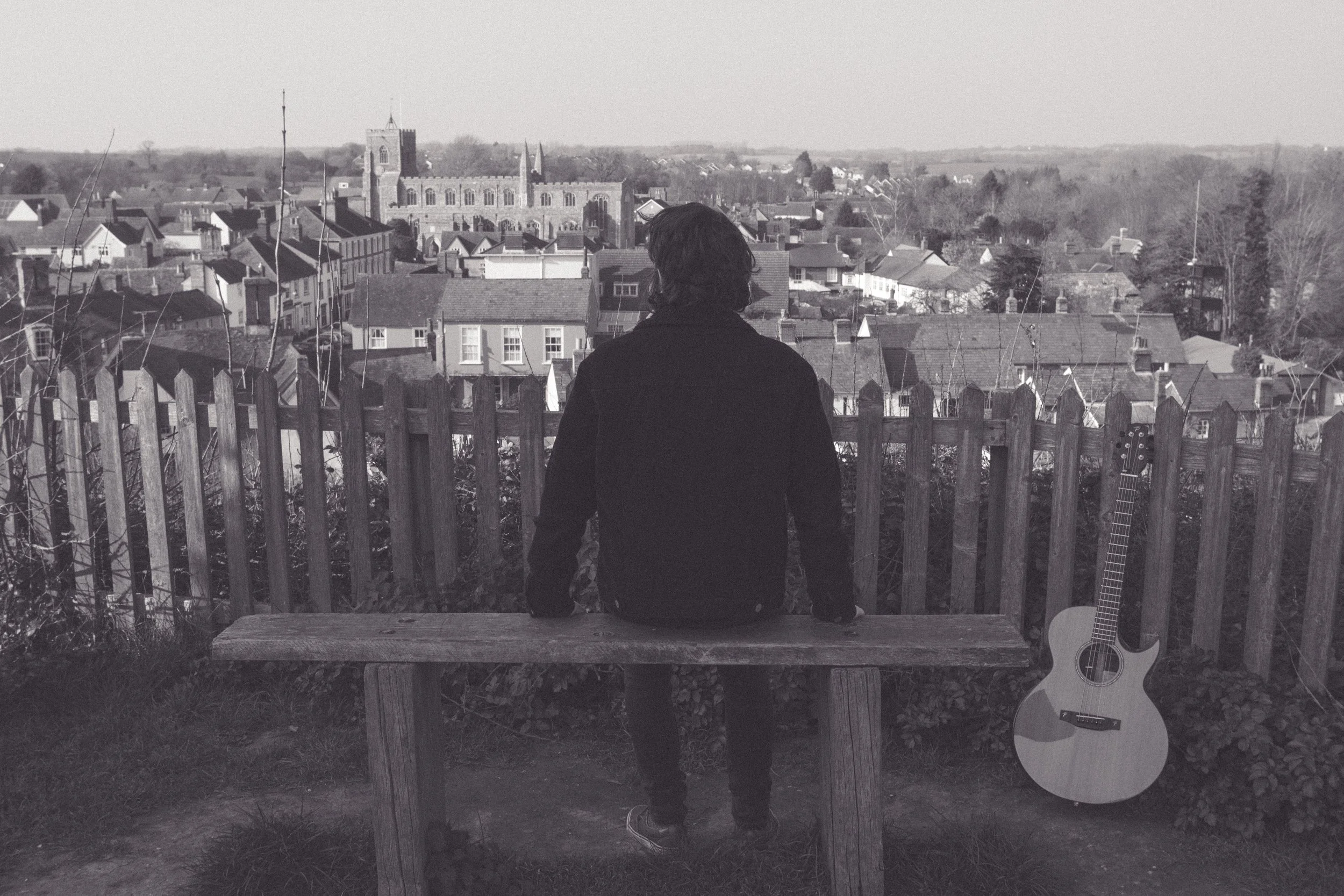 A person with dark, shoulder-length hair wearing a dark jacket and jeans, standing with their back to the camera, on a wooden bench looking over a town with a large church or cathedral in the distance. A guitar is leaning against the fence to the right.