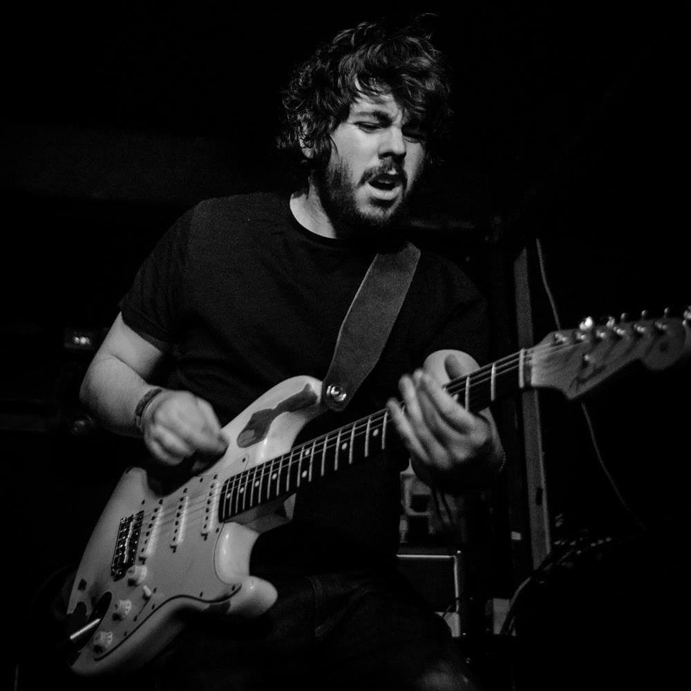 A man with curly hair and a beard (Steve) playing an electric guitar in a dark room.