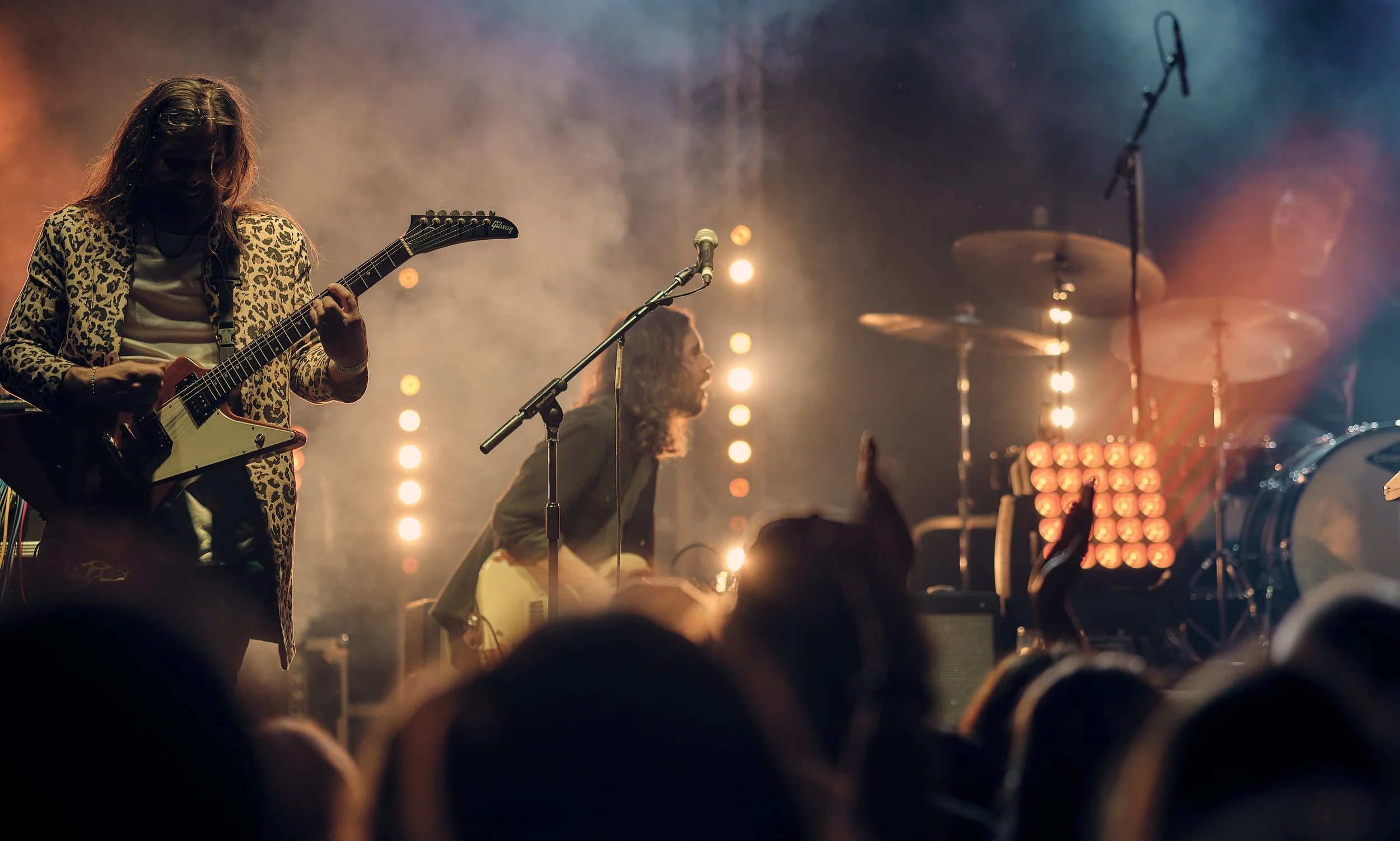 Musicians performing on stage with colorful lighting, one playing an electric guitar and another singing into a microphone, surrounded by an audience.