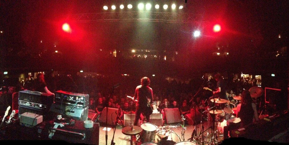 A band performs on stage in a dimly lit venue with a red glow, with the audience watching.