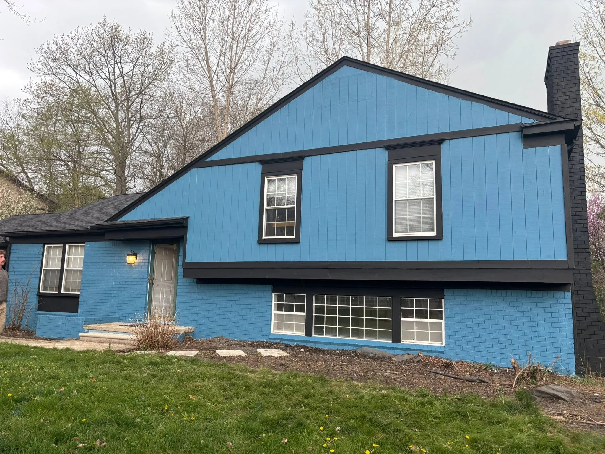 A two-story blue house with black trim and a black brick chimney. The front yard has grass and some small bushes, with a concrete path leading to the front door.