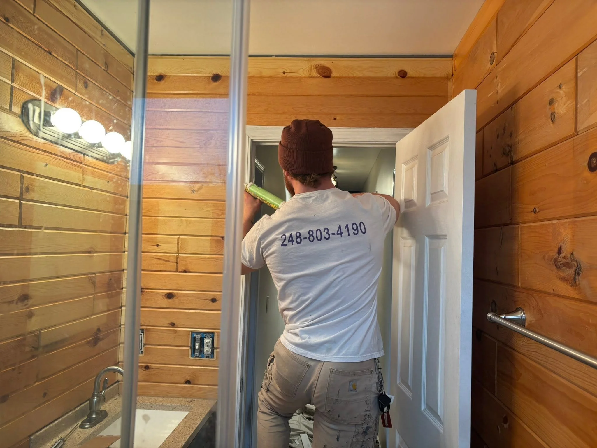 A man wearing a white t-shirt and beige cargo pants is working on electrical wiring inside a bathroom with wood-paneled walls. He is holding a green tool and facing a wall with electrical outlets and switches. The bathroom has a countertop with a sink, a mirror, and a lighting fixture with multiple bulbs.