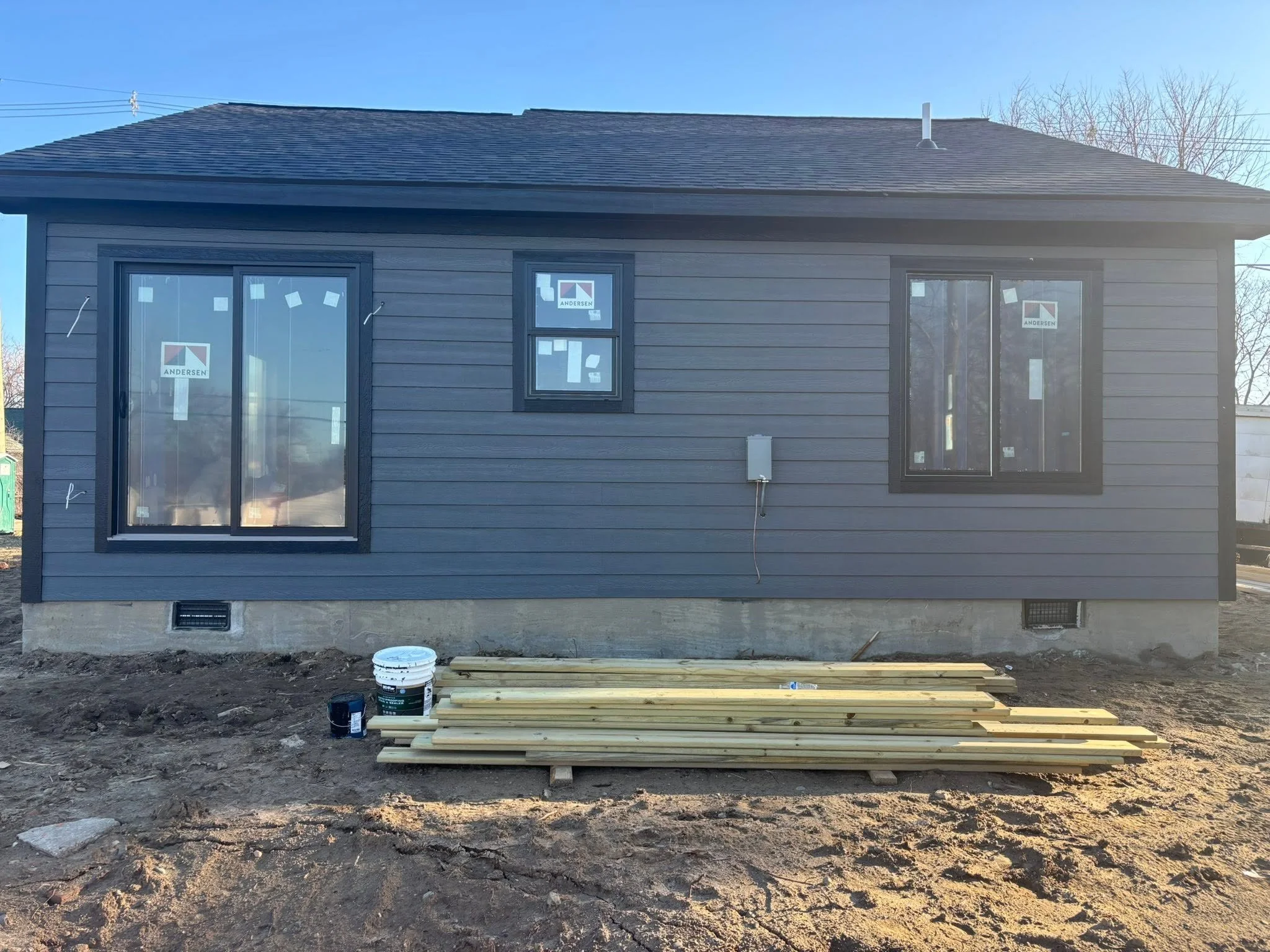 Back exterior of a house under construction with gray siding, multiple windows with stickers, and building materials like wood planks, paint buckets, and a bucket of concrete on the ground.