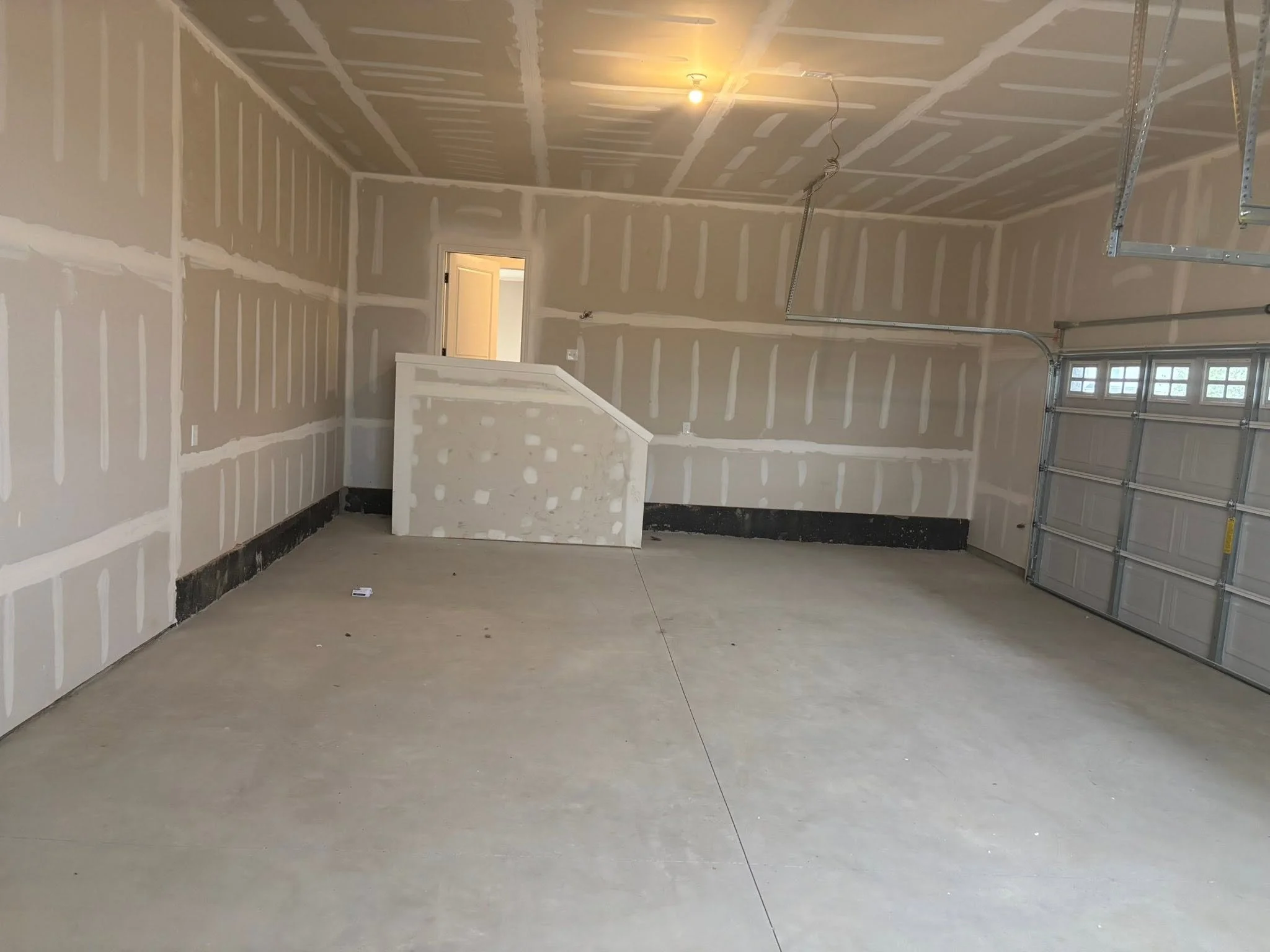 An unfinished garage with drywall installed on the walls and ceiling. The drywall seams have been taped but not yet painted. There is a garage door with windows on the right side and an open door leading to another room on the left. A small section o