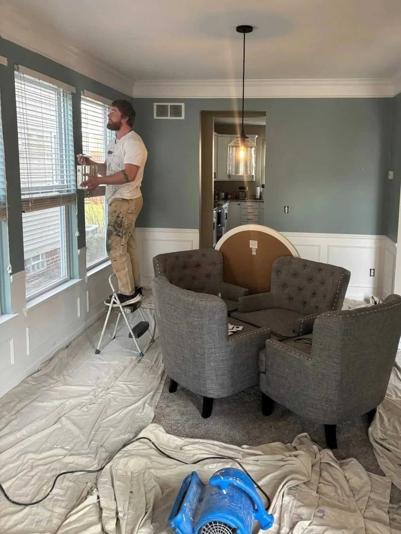 A man standing on a stepladder painting window shutters in a living room while holding a small paint roller and paint can. The room is covered with painter's plastic on the floor and has a group of upholstered chairs, a small kitchen visible through a doorway, and a hanging light fixture.
