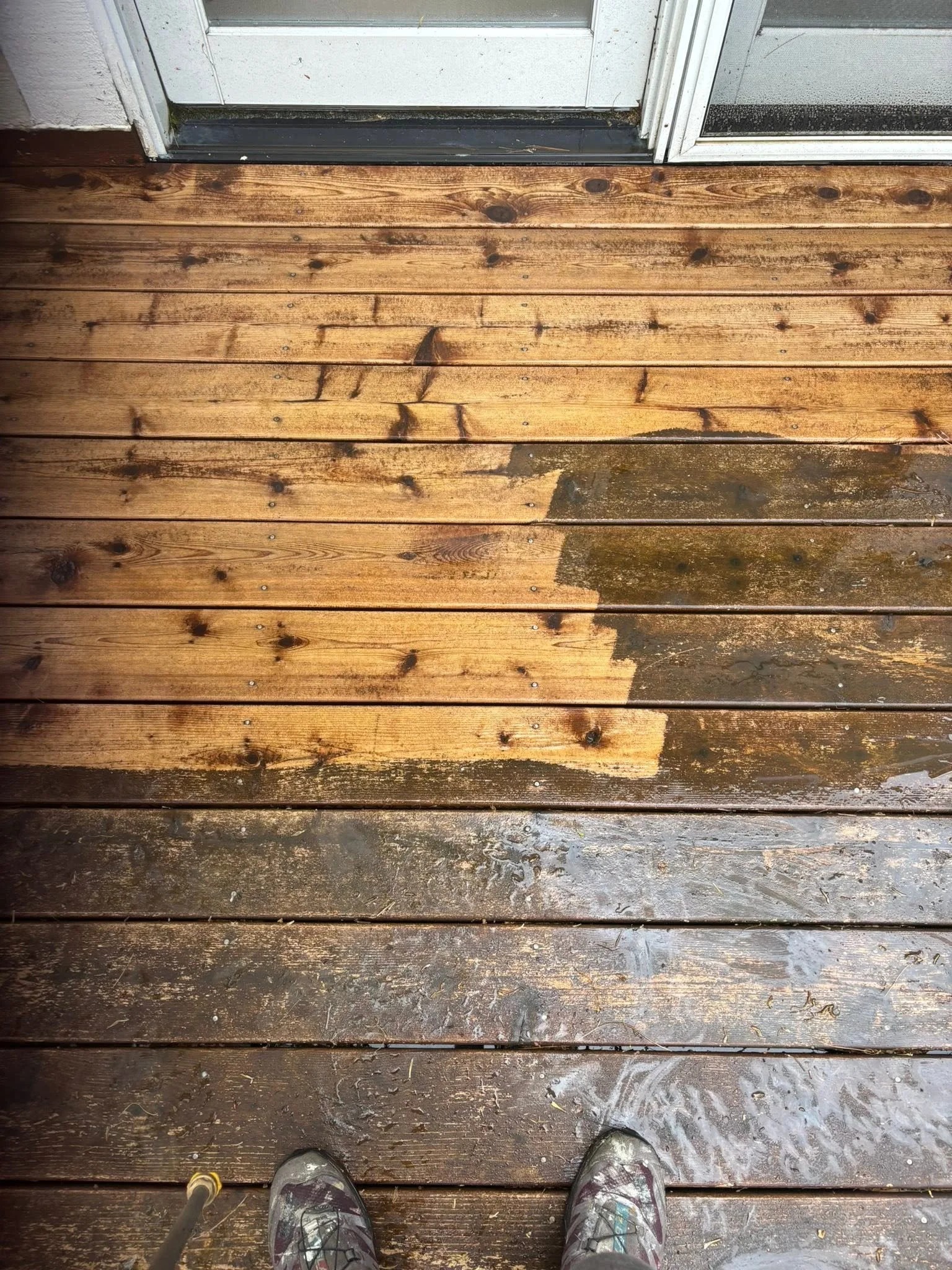 View of a wooden deck with a transition from wet to dry area, as seen from someone's feet wearing sneakers, with a door and screen door in the background.