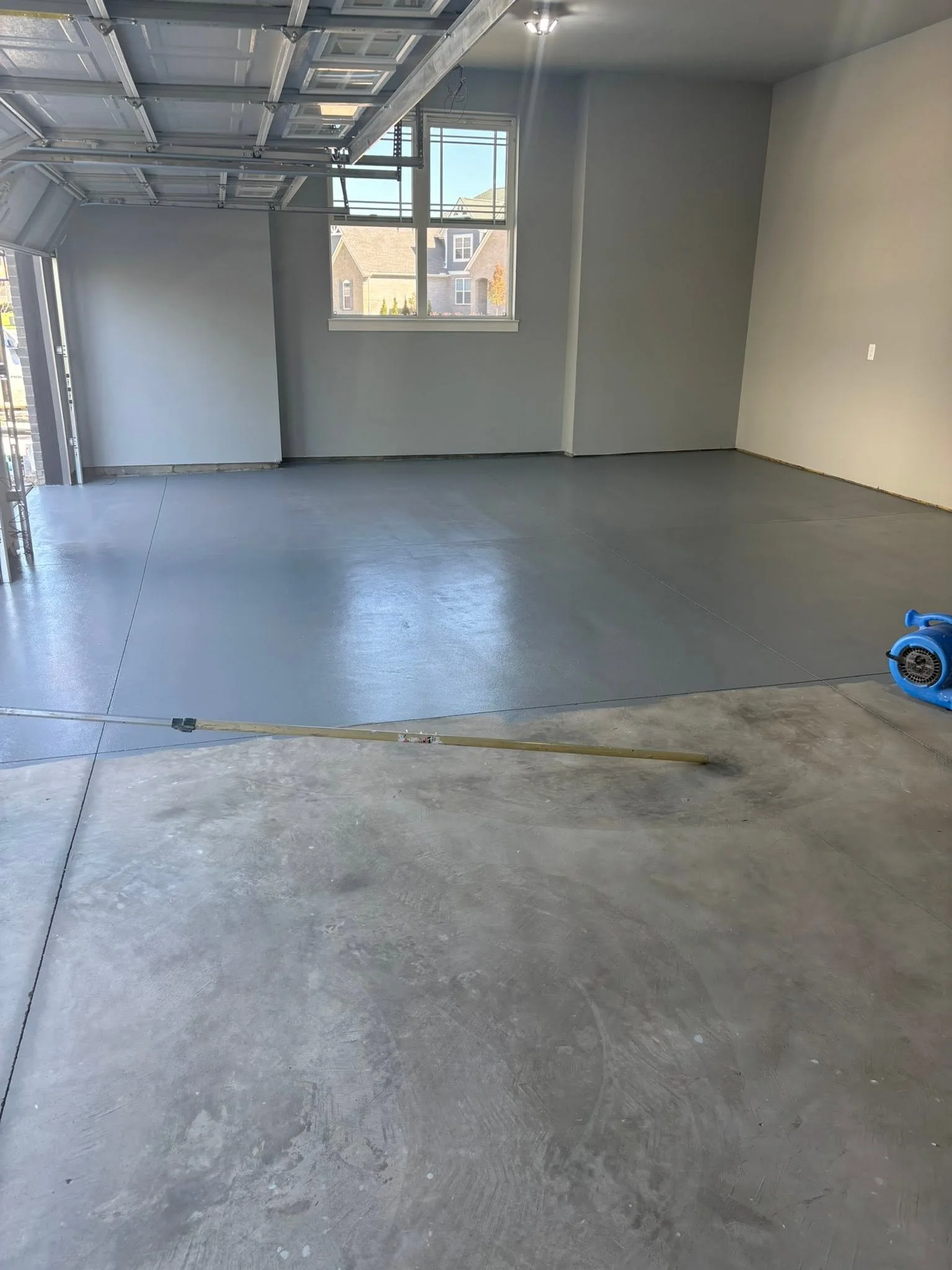 Empty garage with gray concrete floor, white walls, and windows showing houses outside.