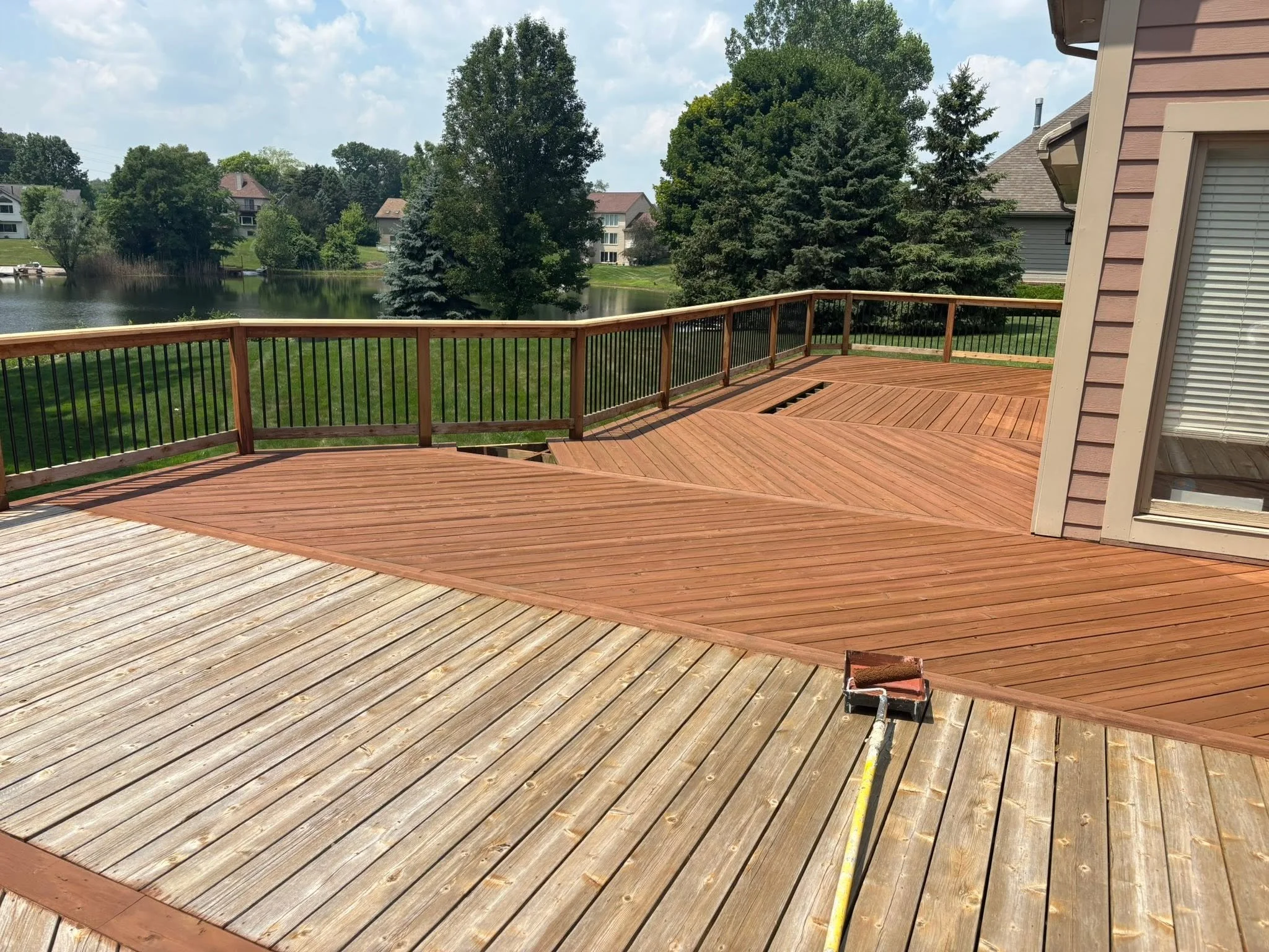 A partially completed wooden deck with a corner railing, a paint roller tray, and a yellow extension cord on the deck surface, overlooking a suburban neighborhood with a lake, trees, and houses.