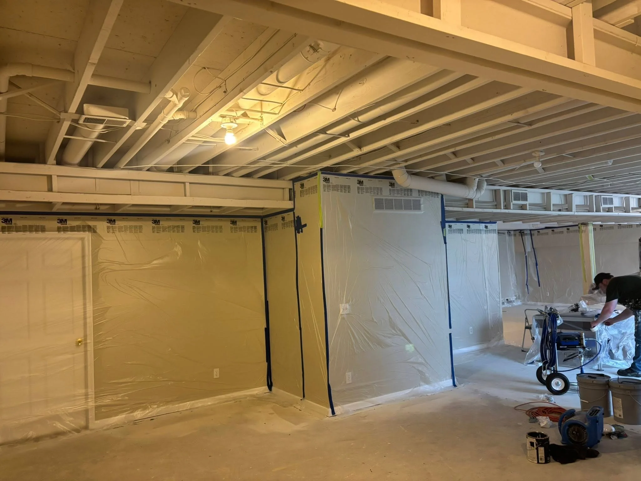 Construction site ceiling with visible pipes, wires, and a light bulb, partially covered with plastic sheeting, with a worker in the background.