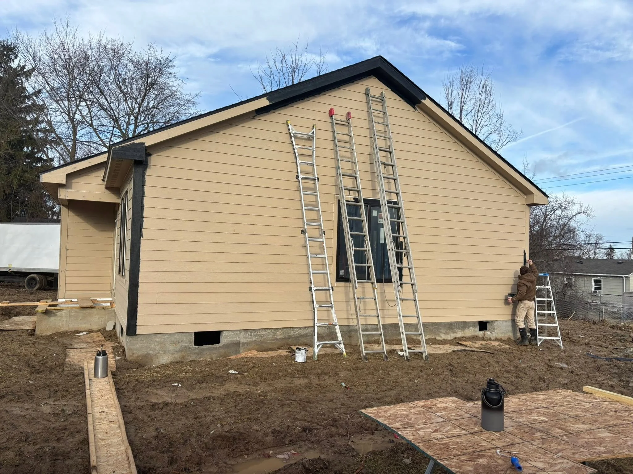 A house under construction with beige siding, multiple ladders leaning against it, and workers working near the foundation. The sky is partly cloudy with leafless trees in the background.