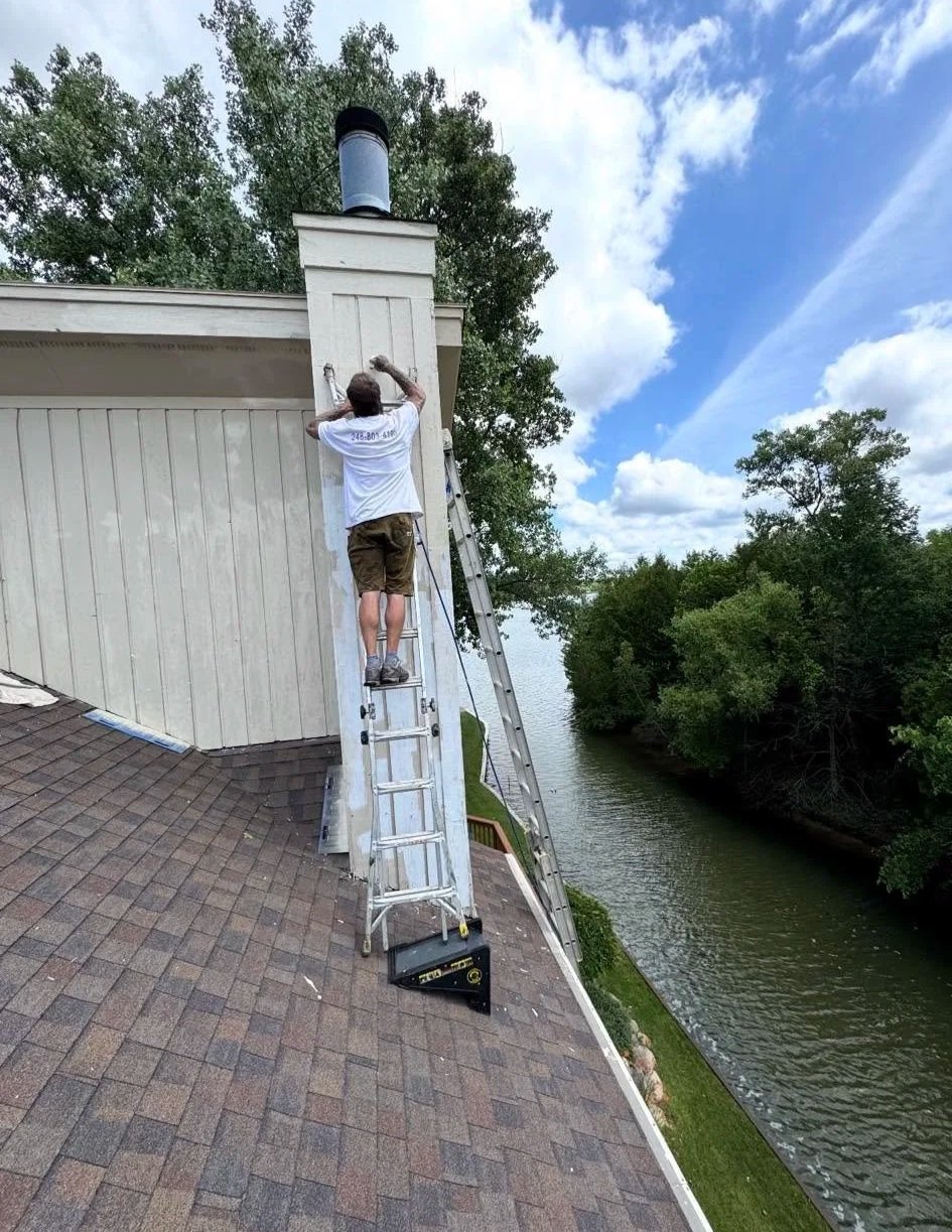 A person is painting the exterior of a house's chimney near the roofline, using a ladder for access, with a river and trees in the background on a partly cloudy day.