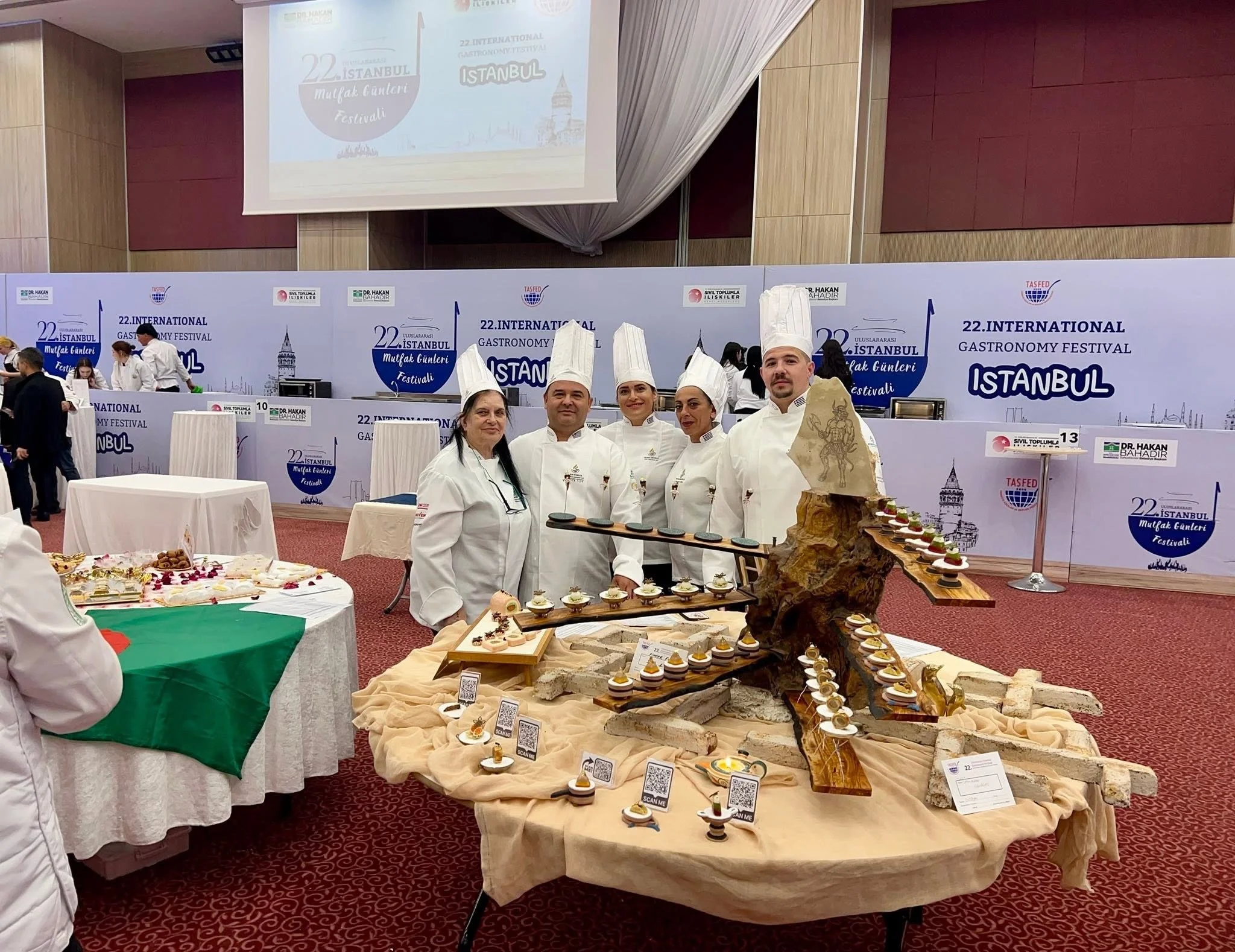 Chefs and participants in a culinary event at the Istanbul International Gastronomy Festival, displaying various prepared dishes and gourmet food items on tables and decorative displays.