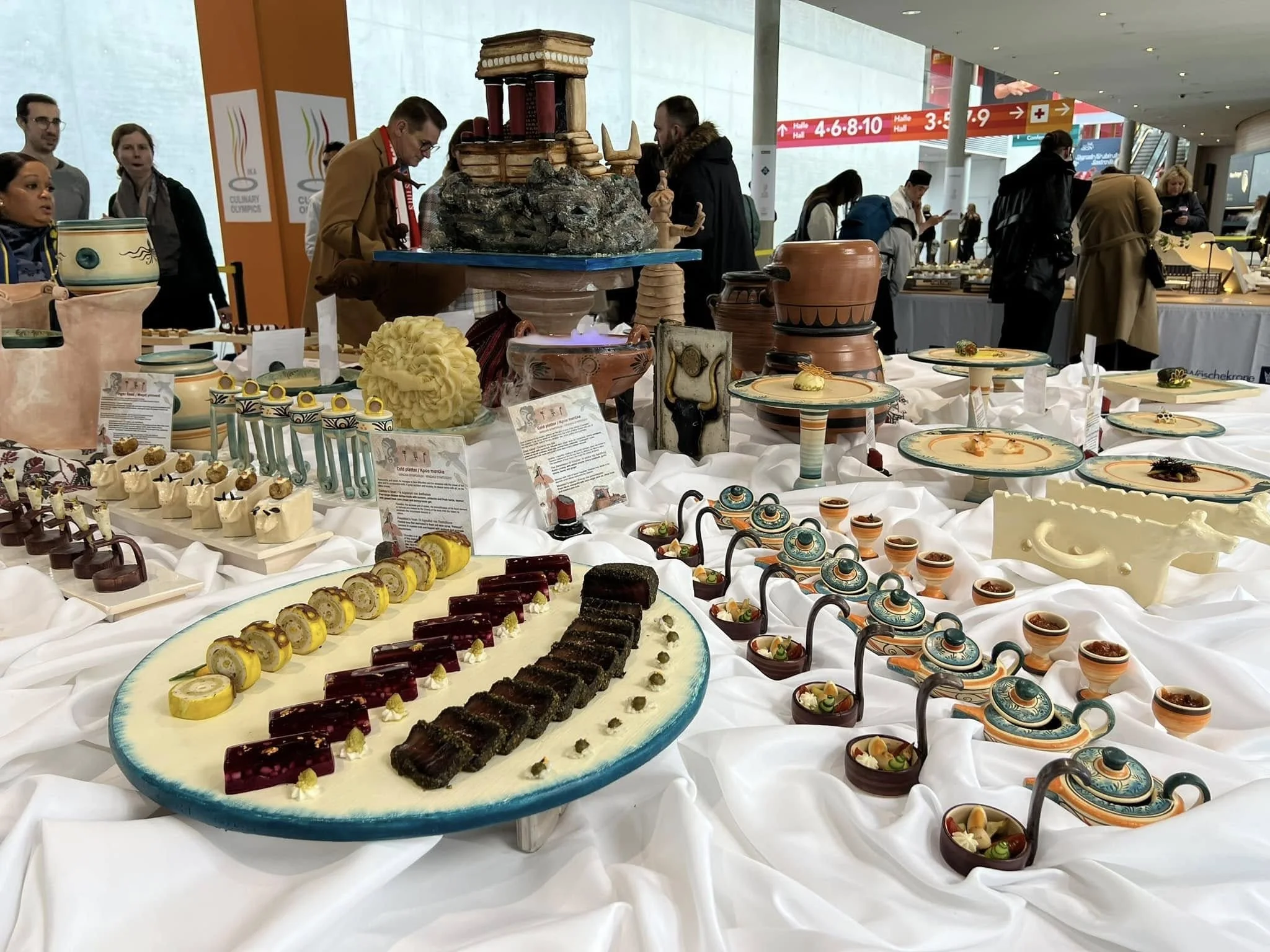 A display of various colorful and intricately decorated pastries, desserts, and pottery at an event in a large, modern indoor space with people browsing in the background.