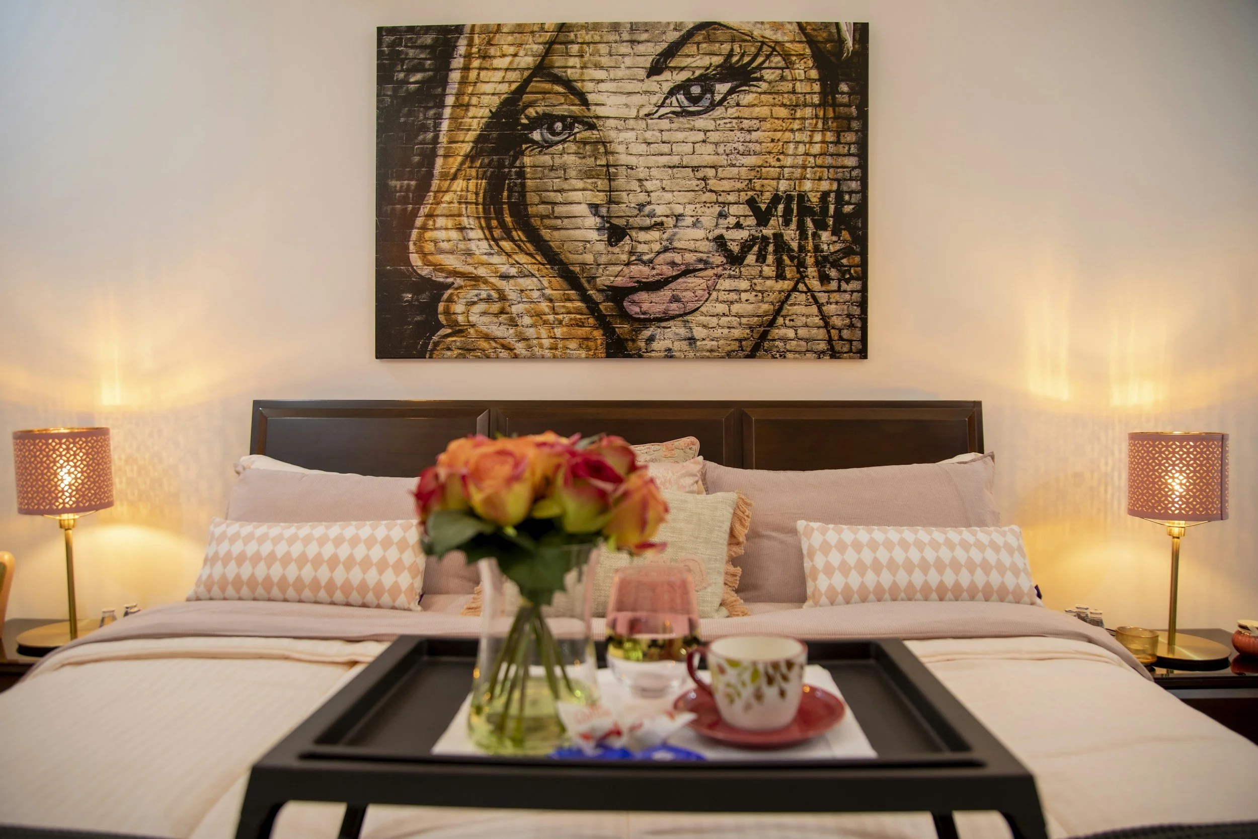 A bedroom with a large painting of a woman's face on the brick wall above the bed. The bed has a beige headboard, pink and white pillows, and a white blanket. There are two matching lamps on nightstands on either side of the bed. A tray on the bed has a flower vase with pink and orange flowers, a cup, and other items.